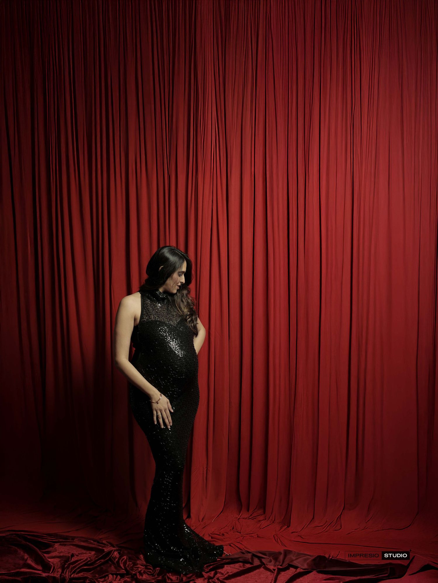 Woman in black sequin gown poses against dramatic red velvet curtain backdrop in elegant studio portrait.