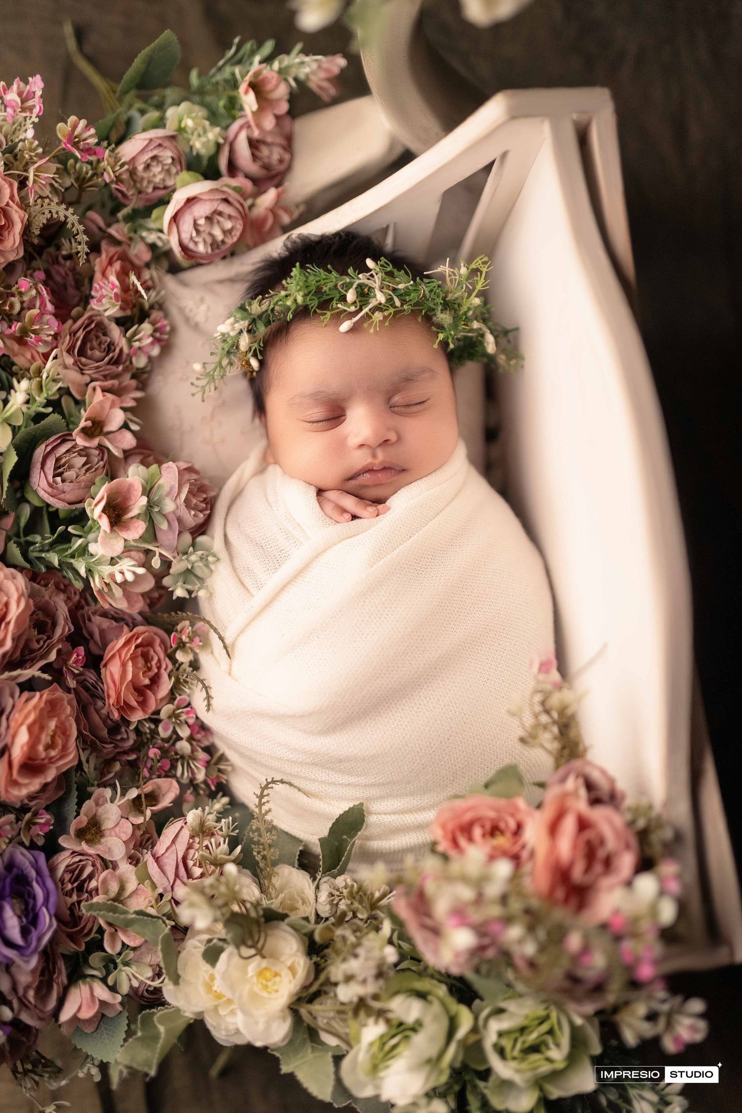 Newborn baby wrapped in white swaddle wearing floral crown, sleeping peacefully surrounded by pink and purple flowers.