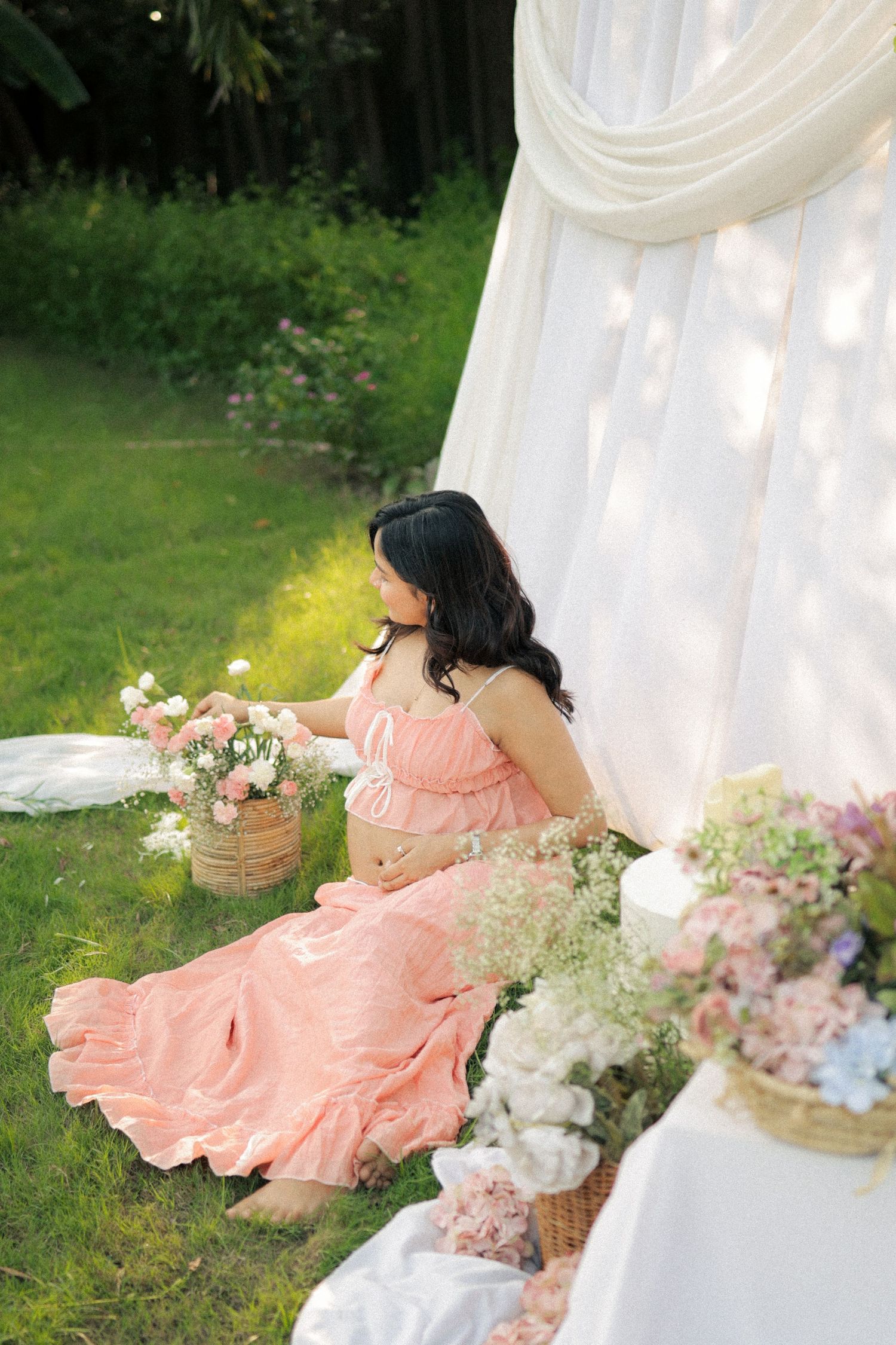 Pregnant woman in pink dress arranging flowers in a basket outdoors near white drapes.