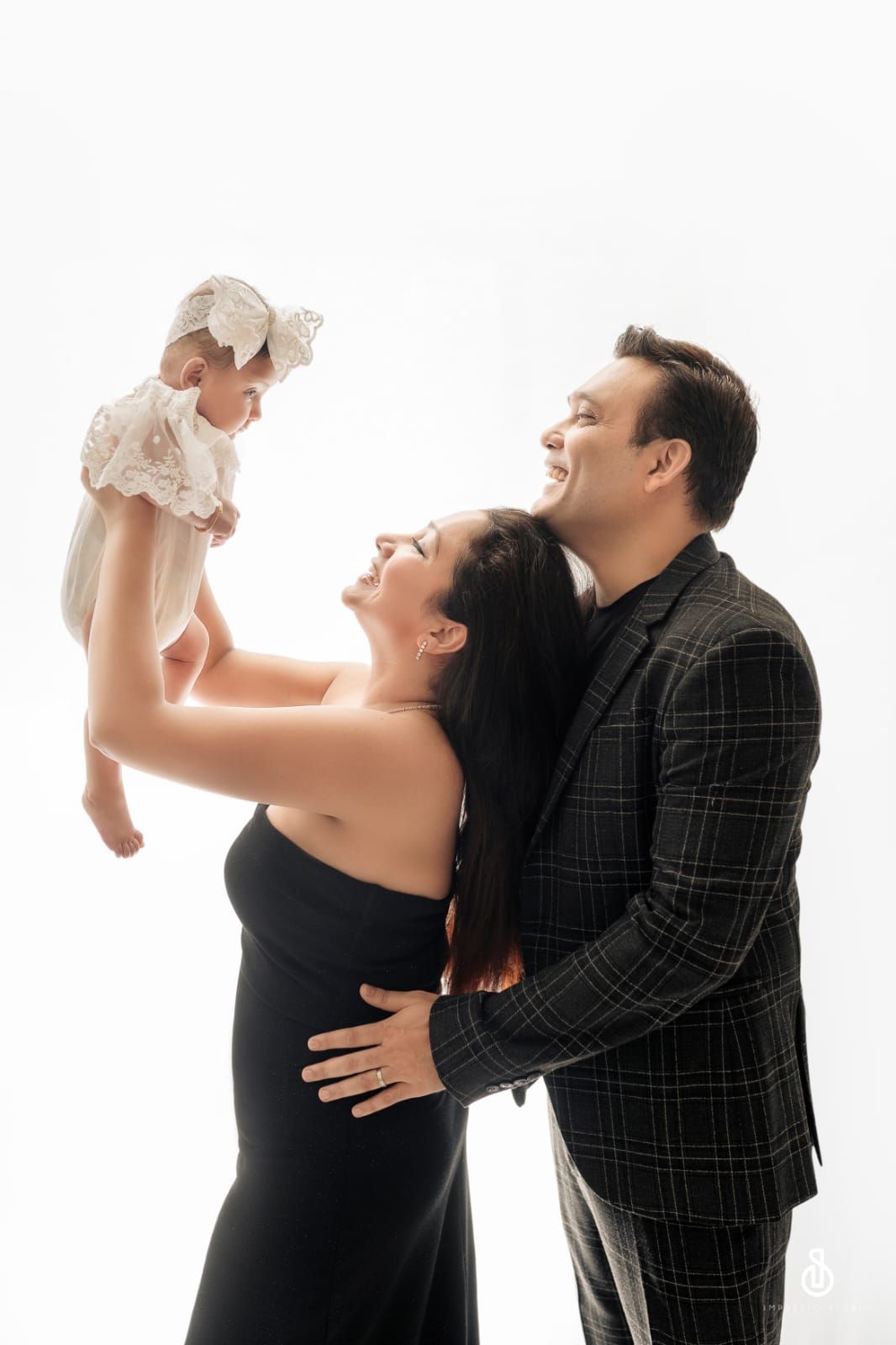 A joyful couple in elegant attire lifts their baby girl in a bright studio portrait.