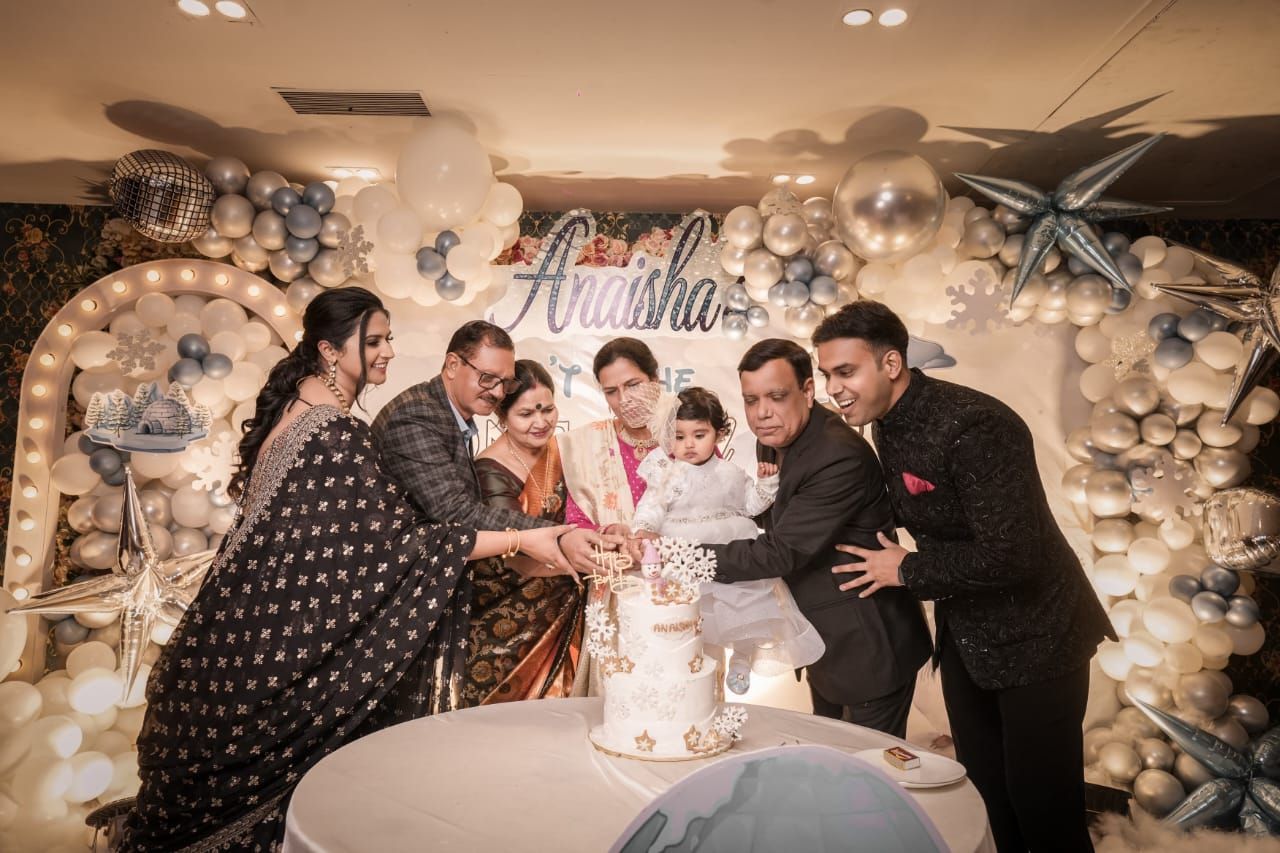 Family celebrating a baby's first birthday party with cake cutting, balloon decorations, and a festive backdrop at an indoor venue.