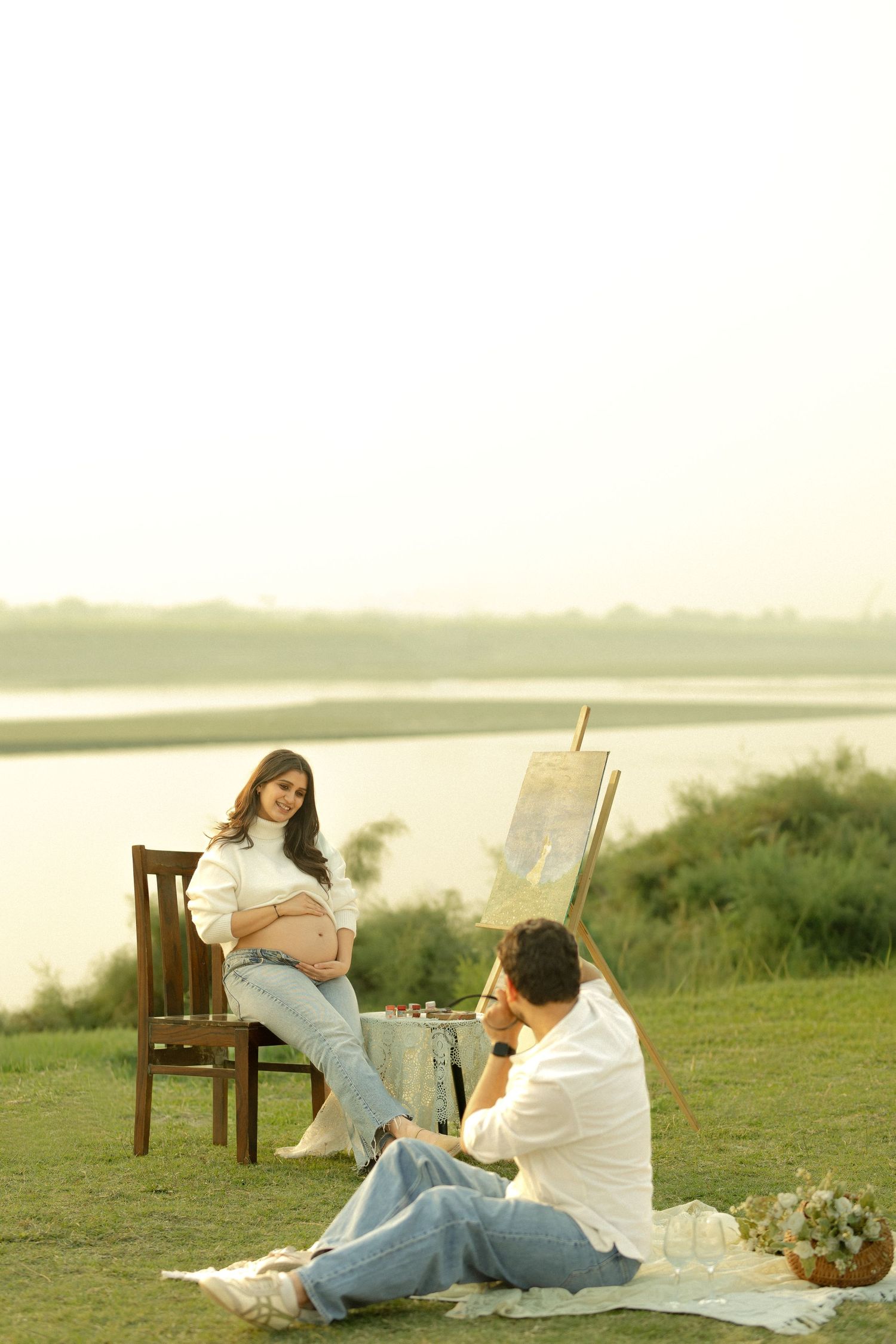 Pregnant woman posing on chair outdoors while partner paints her portrait near a serene lake at sunset.