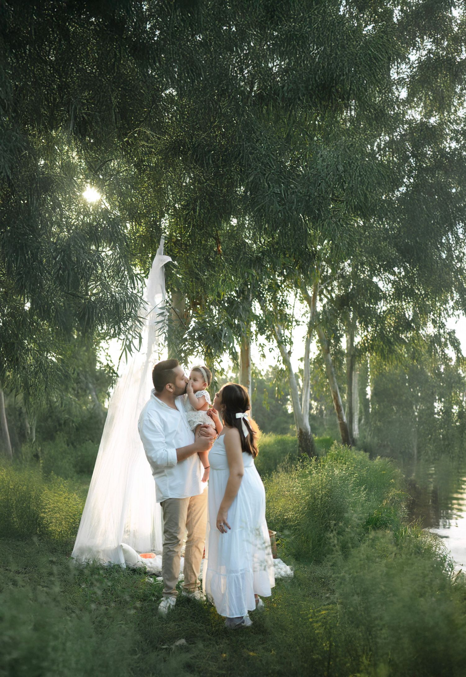 A couple in white outfits shares a romantic moment together by a lake surrounded by trees at sunset.
