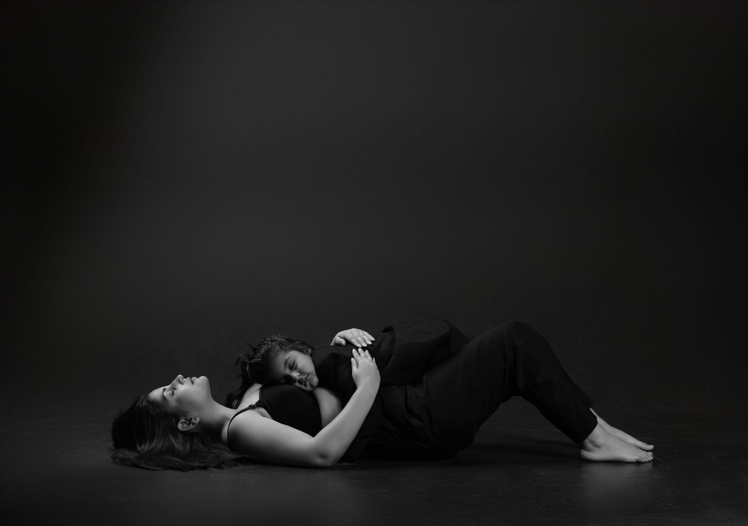 Two people in black clothing lying together on a dark studio floor in a black and white artistic photograph.