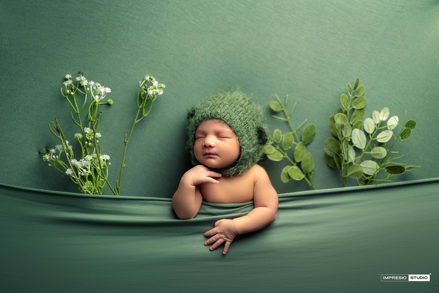 Newborn baby in green knit hat posed among white flowers and greenery on a soft green fabric backdrop.
