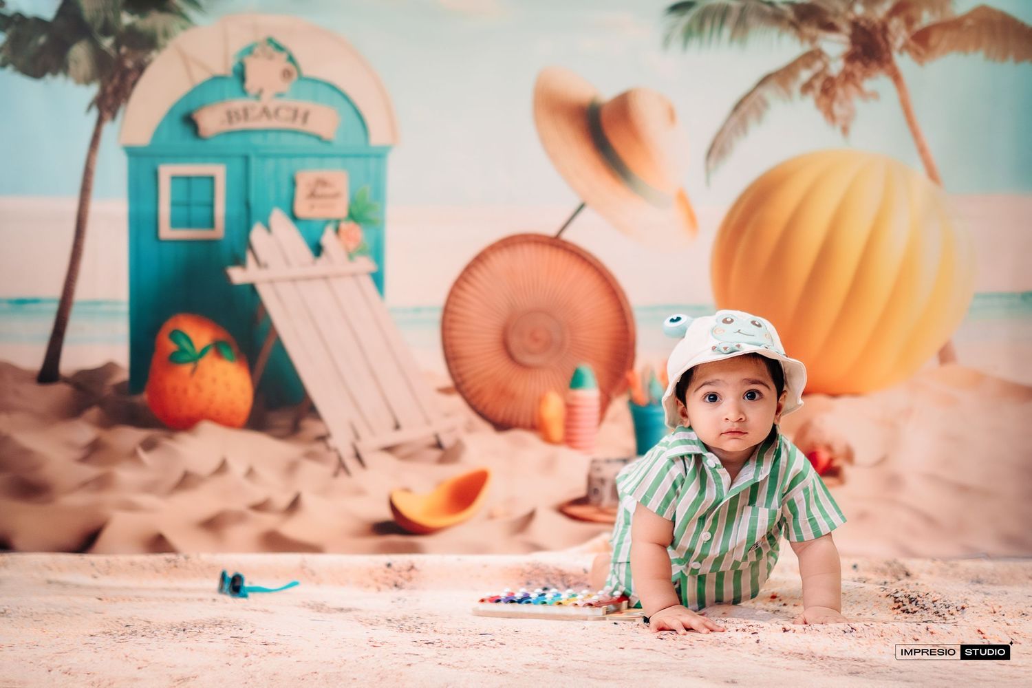 Beach themed baby photo setup with pumpkins, palm trees, and a blue beach house backdrop in soft pastel tones.