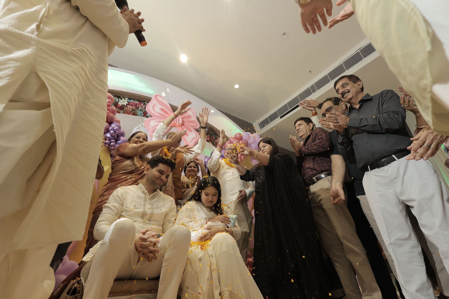 Guests in traditional attire gather around a seated couple in white outfits during a festive Indian wedding ceremony in a decorated hall.