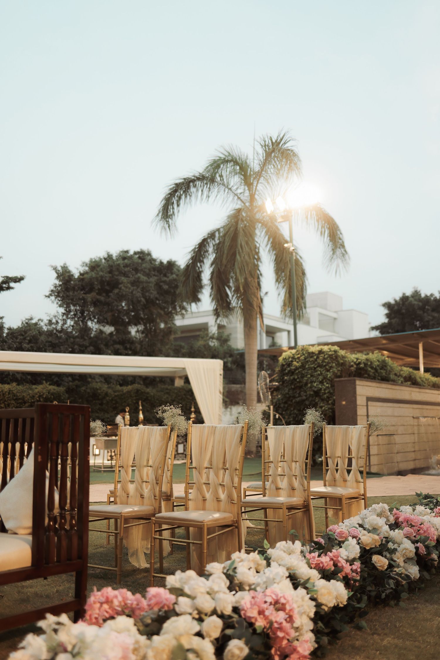 Elegant outdoor wedding ceremony setup with gold chiavari chairs, pink and white floral aisle, and palm tree backdrop.