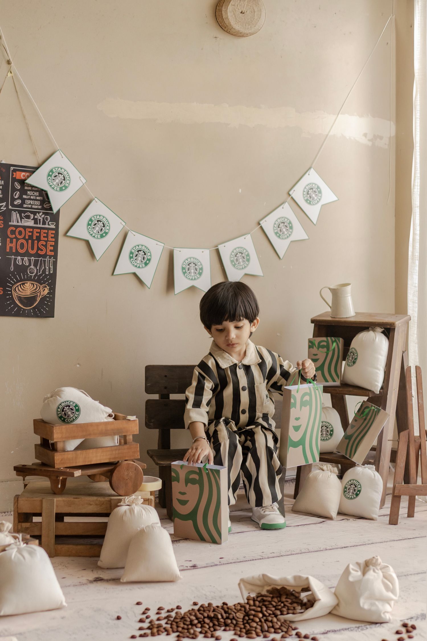 Young child in striped outfit surrounded by Starbucks-themed decor, bags, and coffee beans in a cozy styled photo setup.