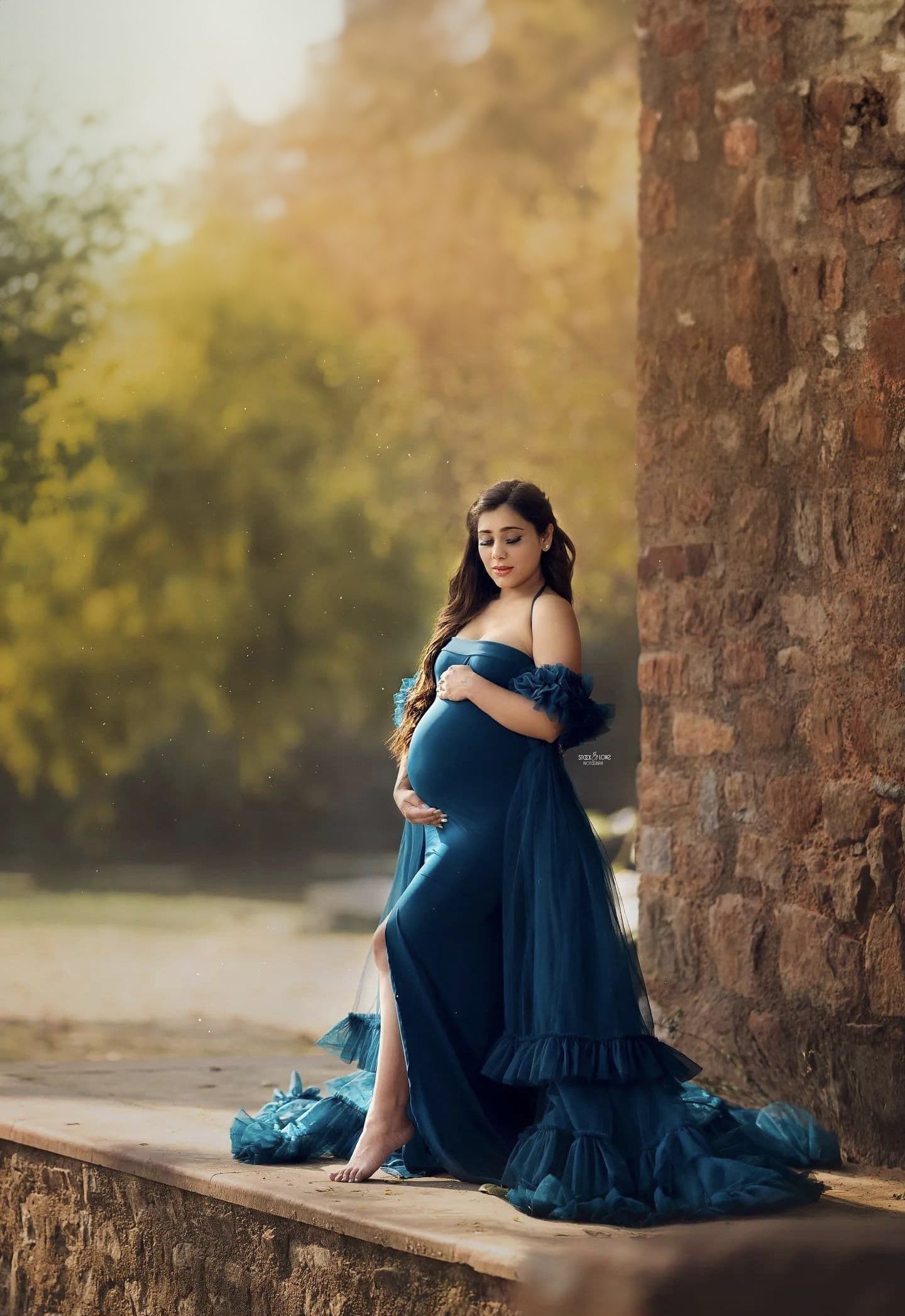 Outdoor maternity photo in a blue flowing gown with woman standing on stone ledge among autumn foliage.