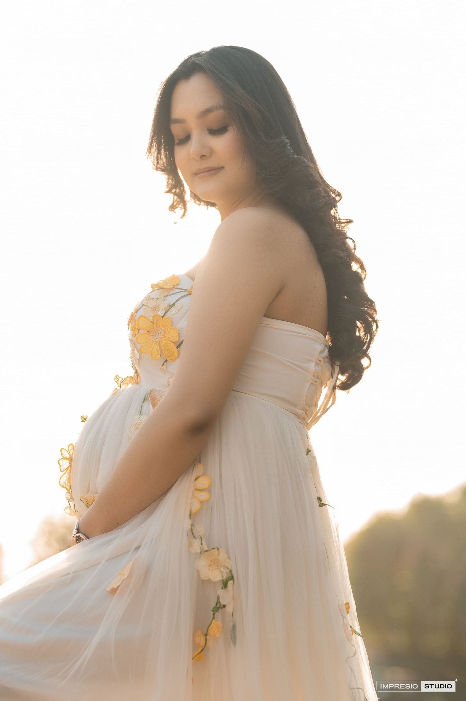 Ethereal maternity portrait in a white dress with yellow floral details captured in golden sunlight.