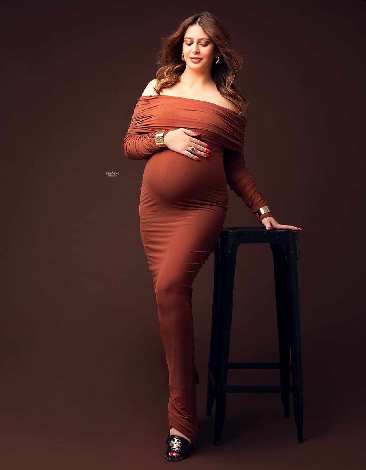Maternity photoshoot featuring rust-colored off-shoulder dress against dark background.
