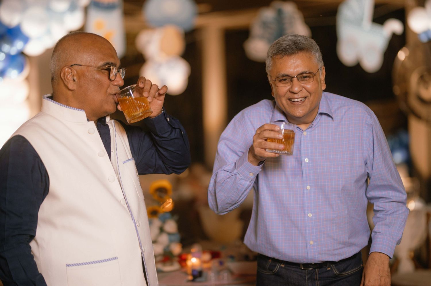 Two men smiling and enjoying drinks together at a festive indoor celebration with decorations in the background.