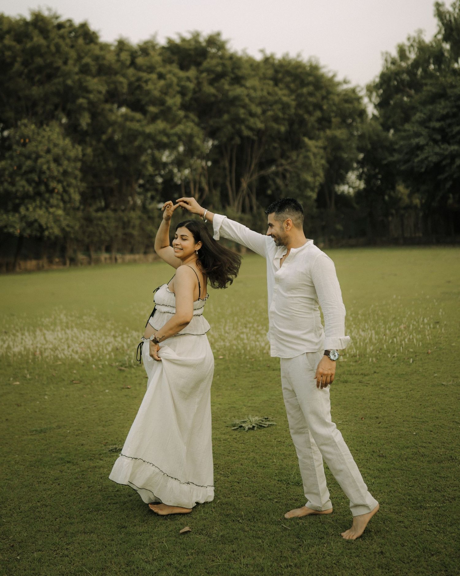 A couple dances together on a green lawn, both wearing white outfits, surrounded by lush trees in a serene outdoor setting.