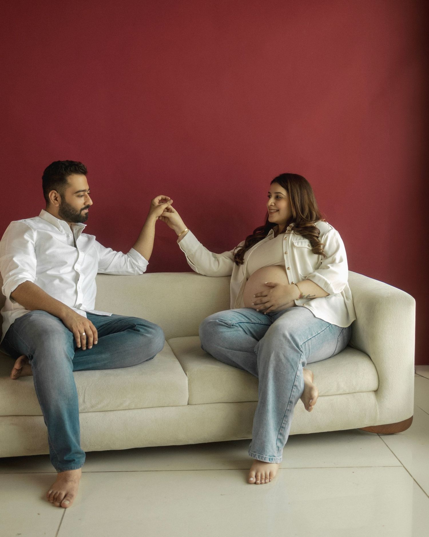 Couple in white shirts and jeans sitting on cream sofa, pregnant woman holding partner's hand against red wall.