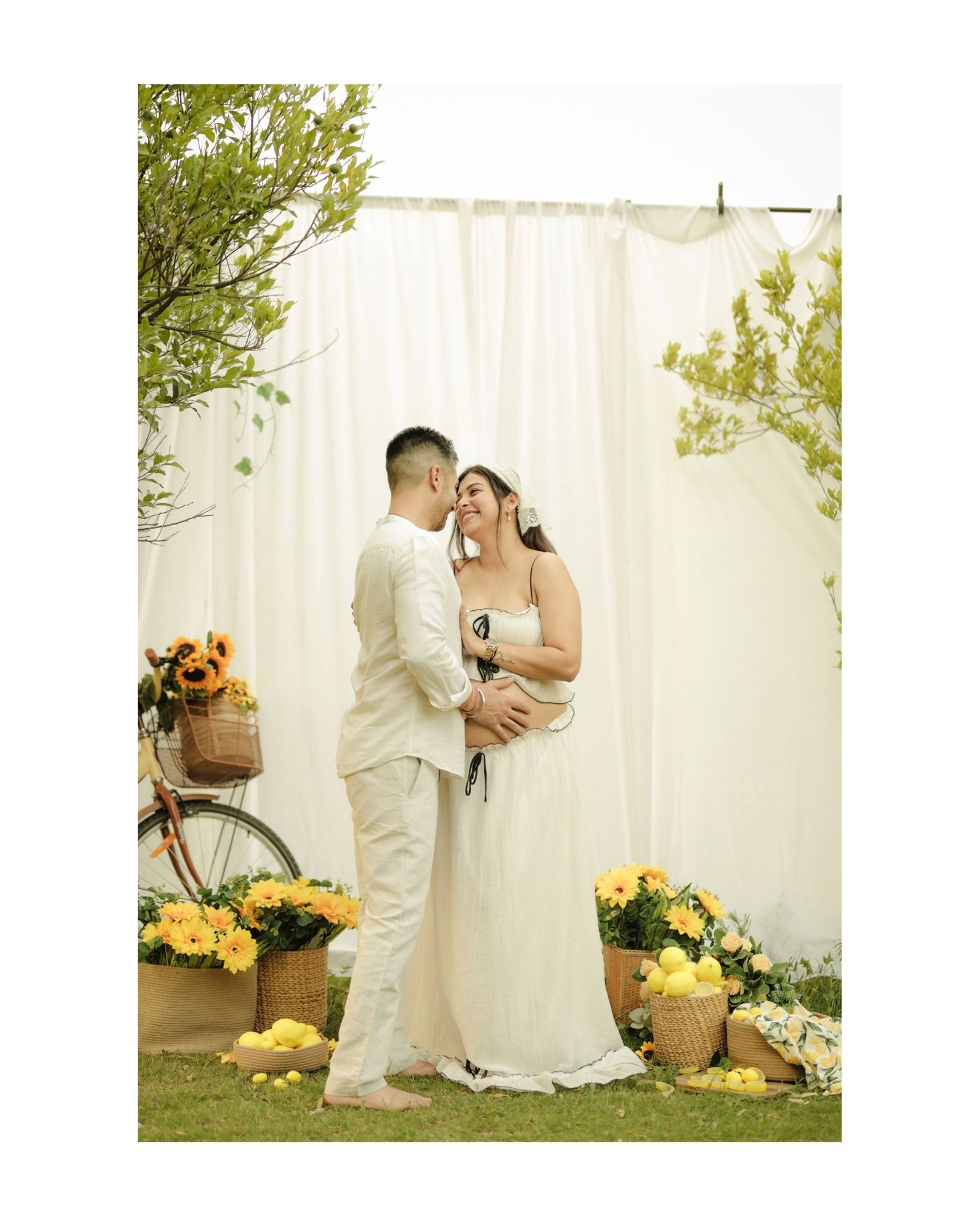 Couple in white attire sharing a romantic moment outdoors with yellow flowers and a white curtain backdrop.