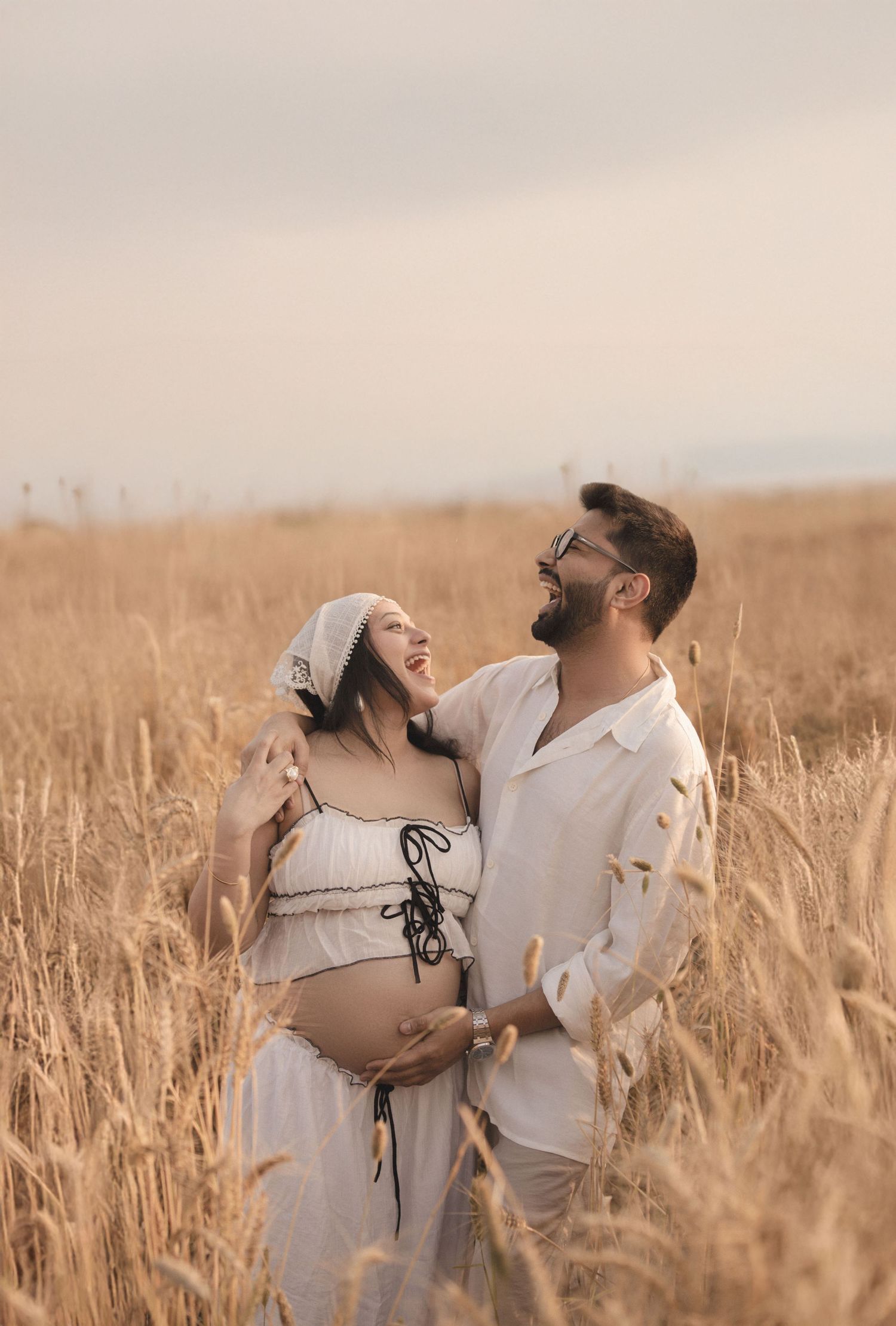 Couple in white outfits embracing in golden wheat field, pregnant woman smiling up at bearded man during maternity photoshoot.
