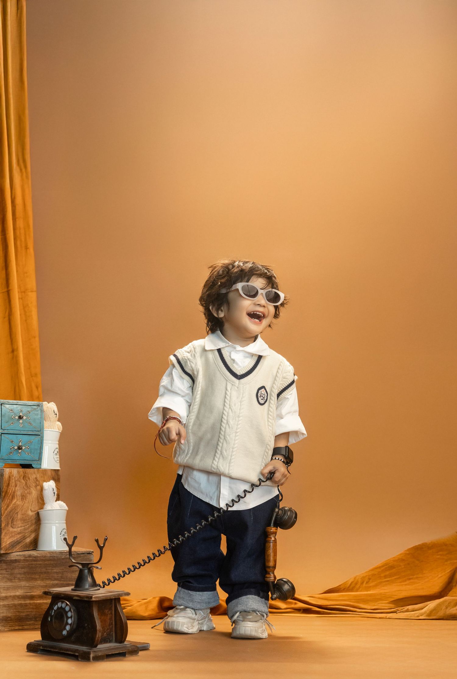 Adorable toddler in white vest, sunglasses, holding vintage phone cord, posing against warm orange backdrop.