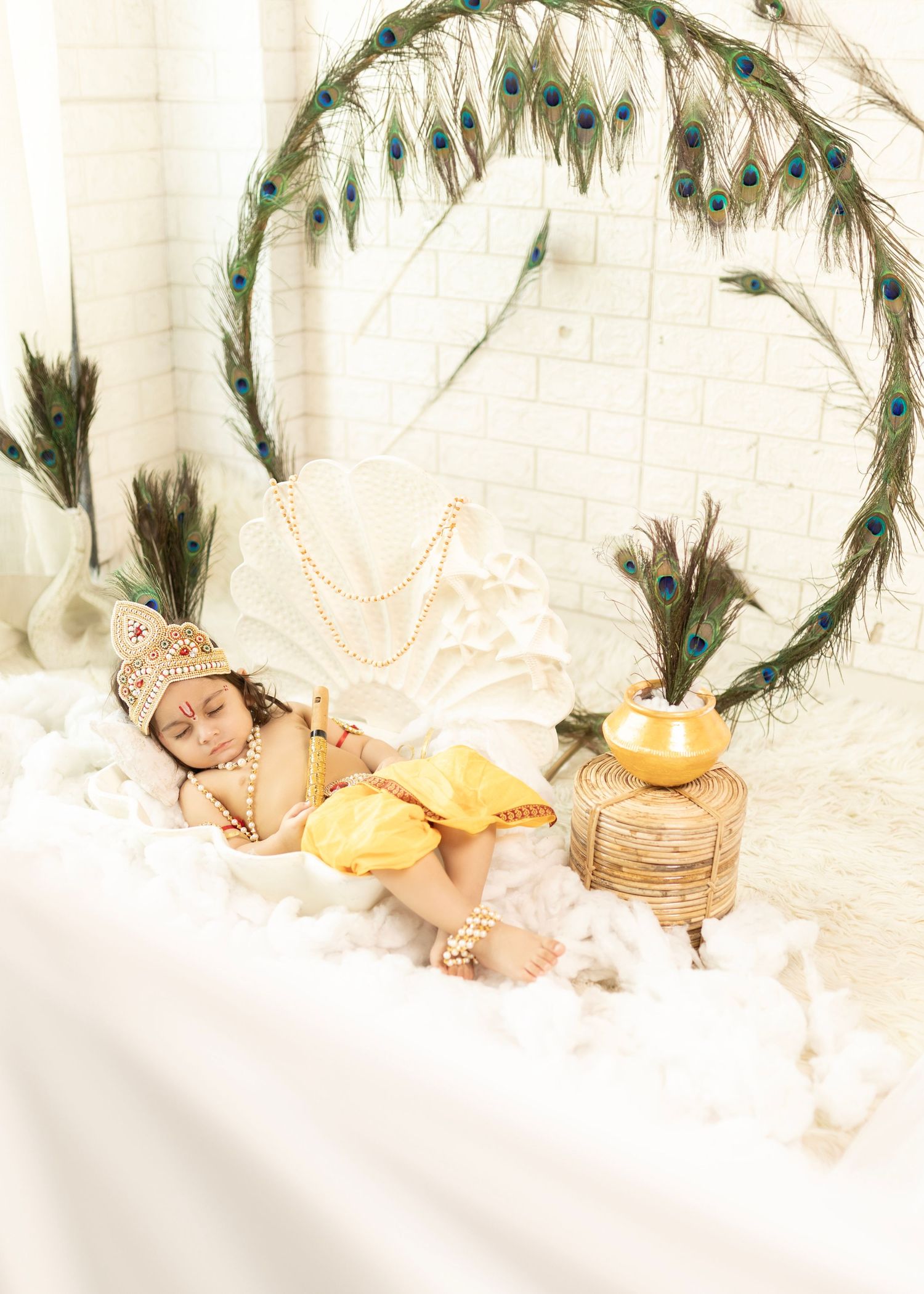 Baby dressed as Lord Krishna in yellow outfit, sleeping peacefully amid peacock feathers and decorative props.