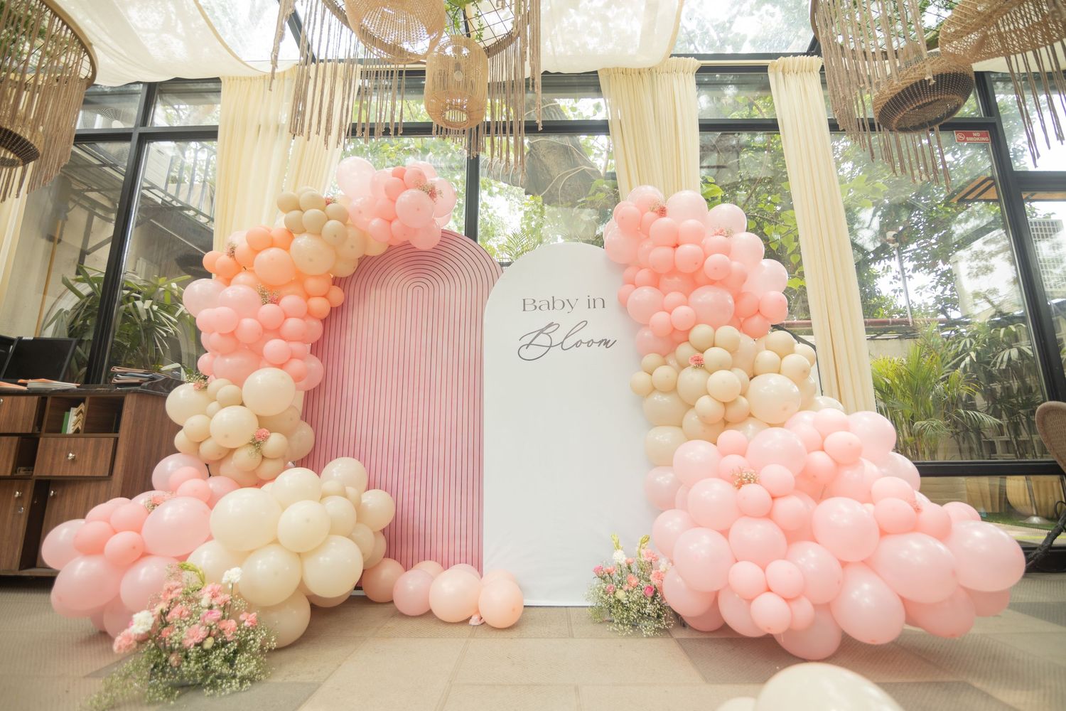 Pink and white balloon arch with floral decorations framing an elegant event backdrop with pastel fringe curtain.