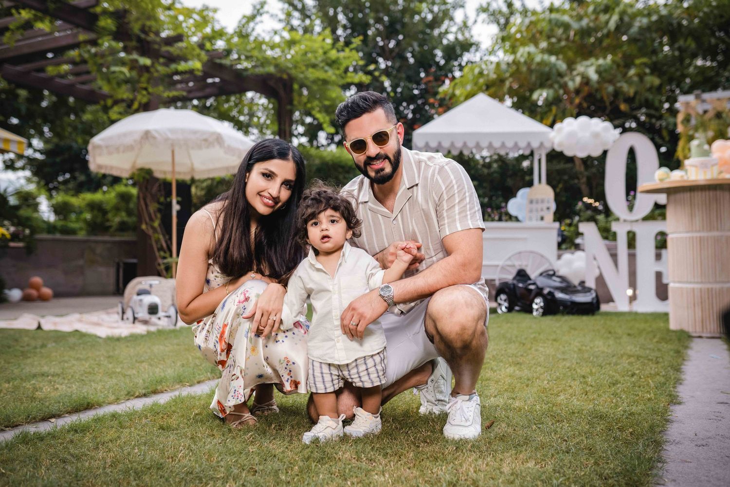 A smiling family of three poses outdoors on green grass, with parents crouching beside their toddler at a garden celebration.