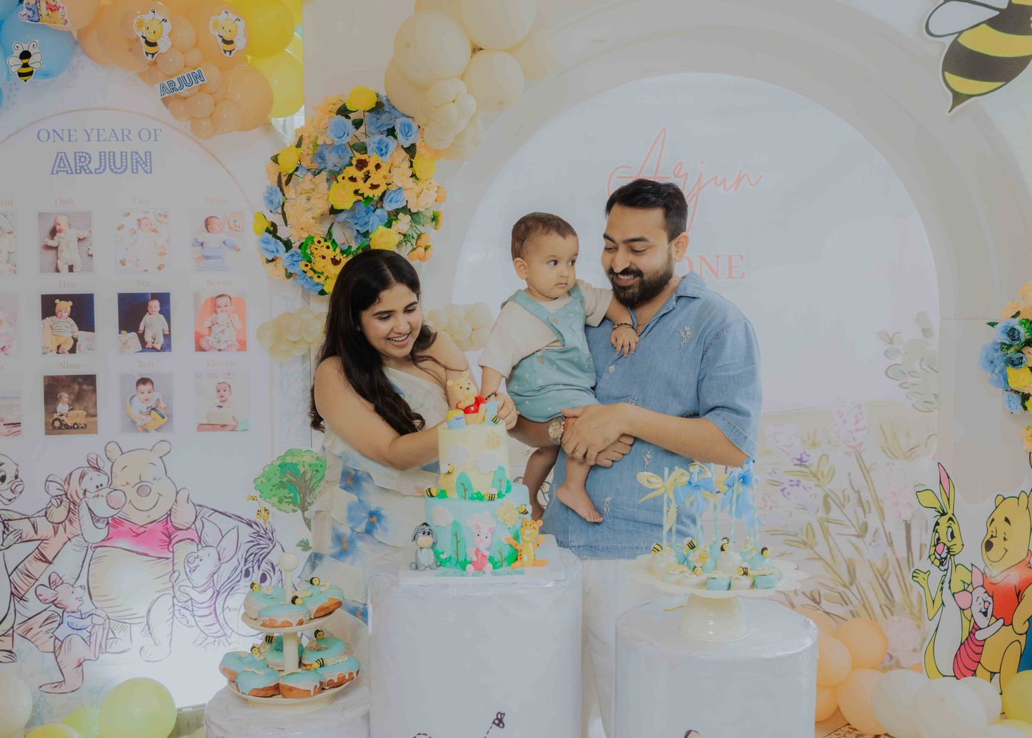 Family celebrating baby Arjun's first birthday with a Winnie the Pooh themed cake and colorful balloon decorations.