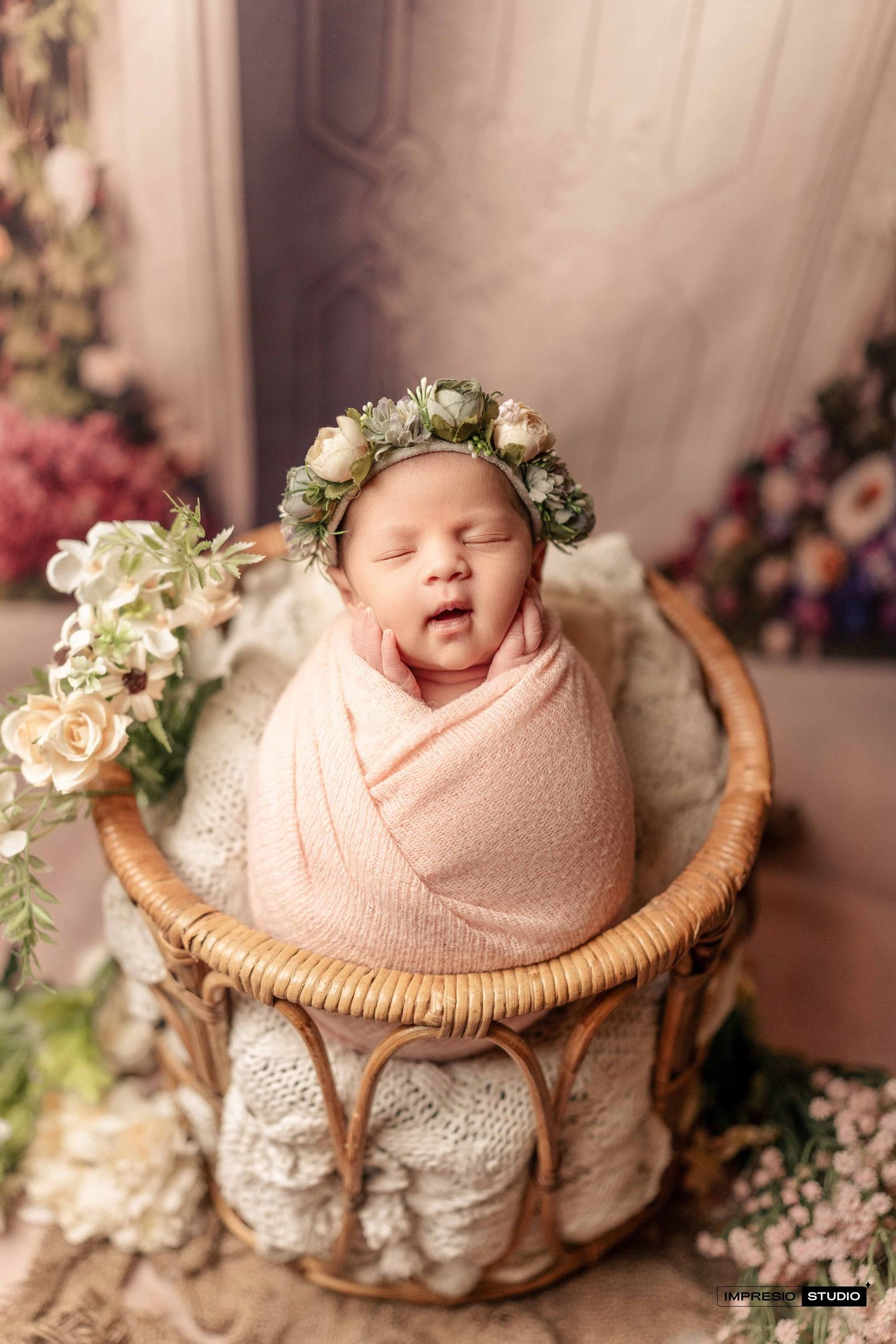 Newborn baby wrapped in pink swaddle wearing floral crown sleeping in wicker basket with flowers.