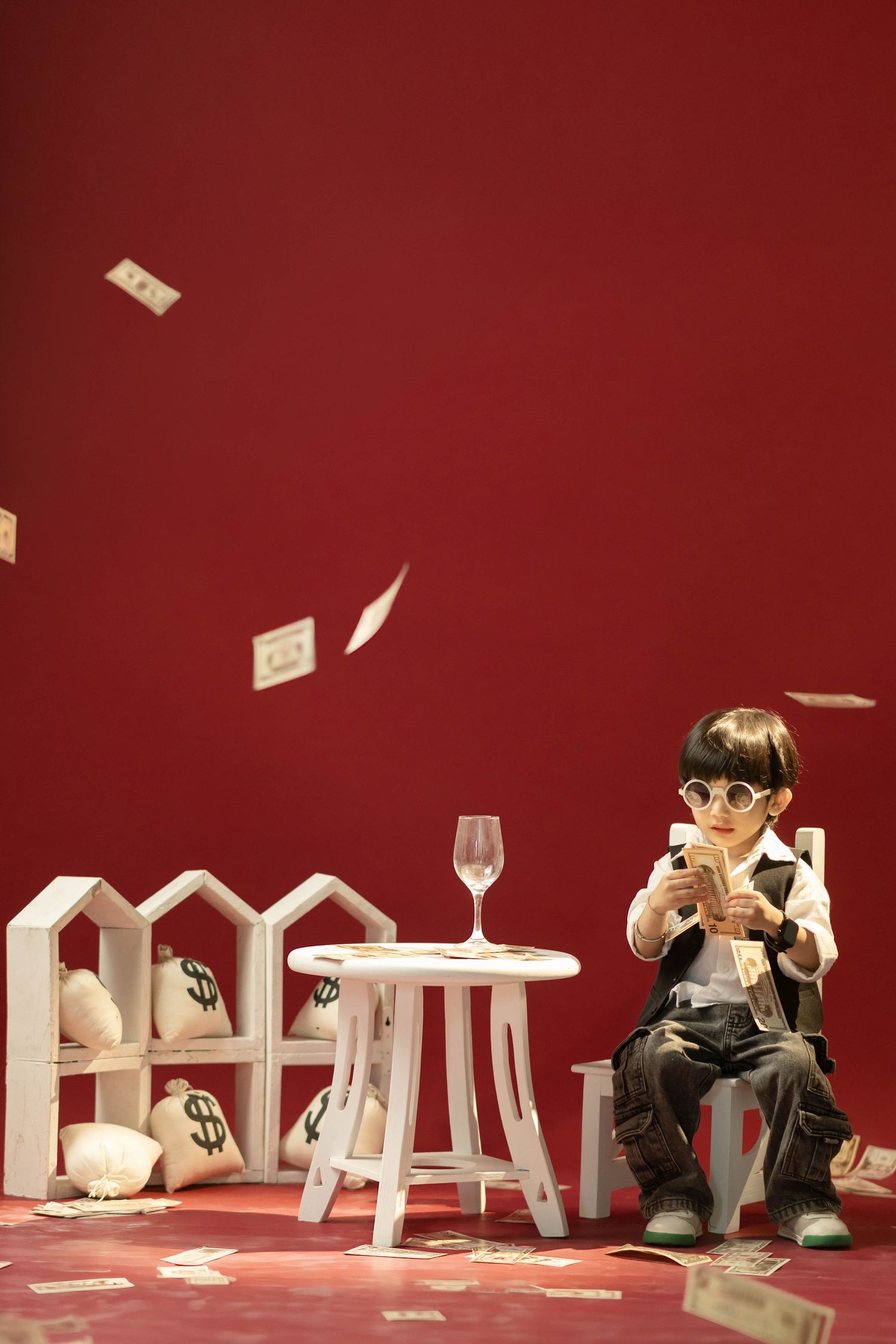 A child in sunglasses sits counting money with bags of cash nearby as dollar bills rain down on a red background.