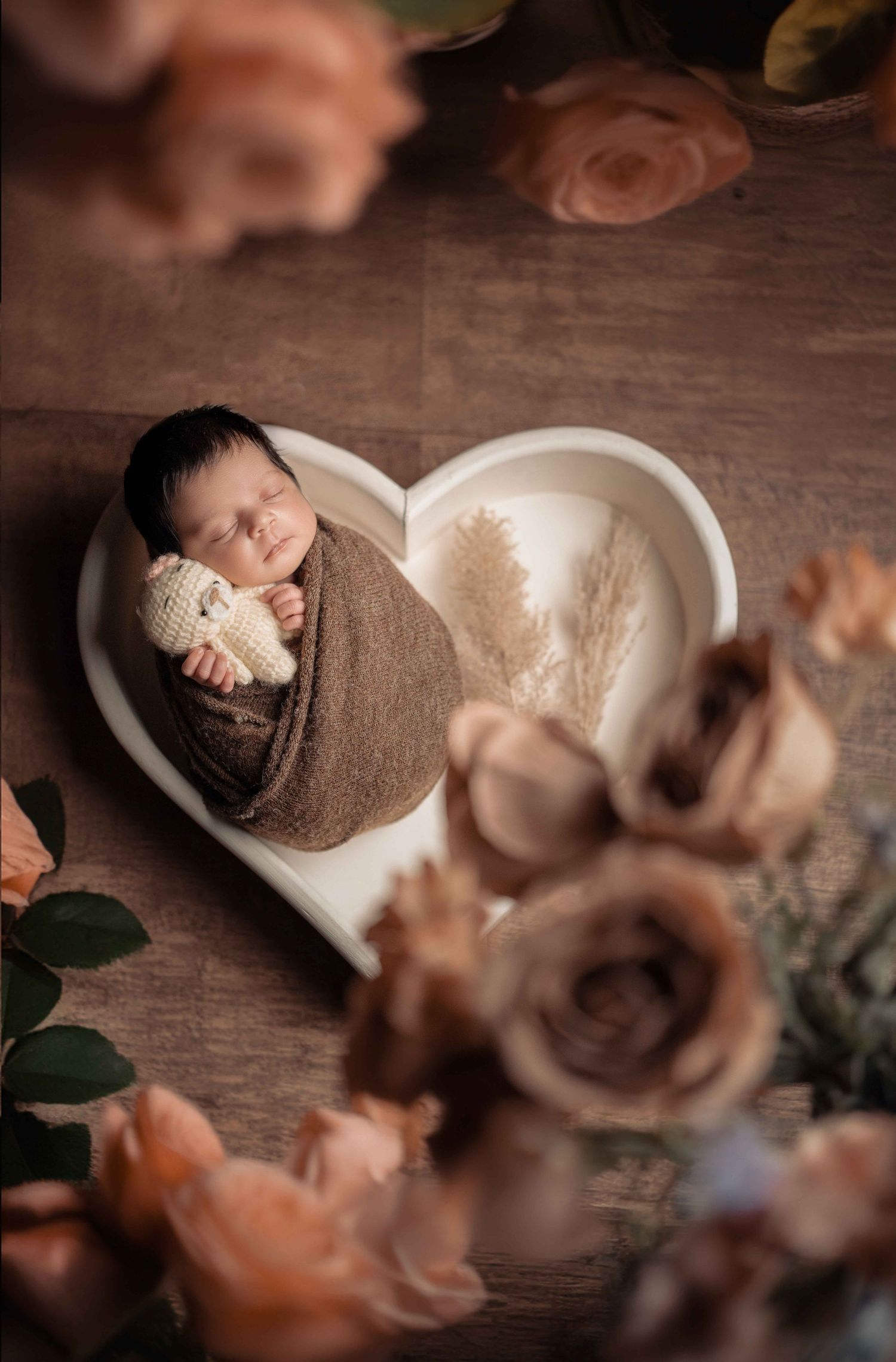 Newborn baby swaddled in brown wrap sleeping in a white heart-shaped bowl surrounded by peach roses.
