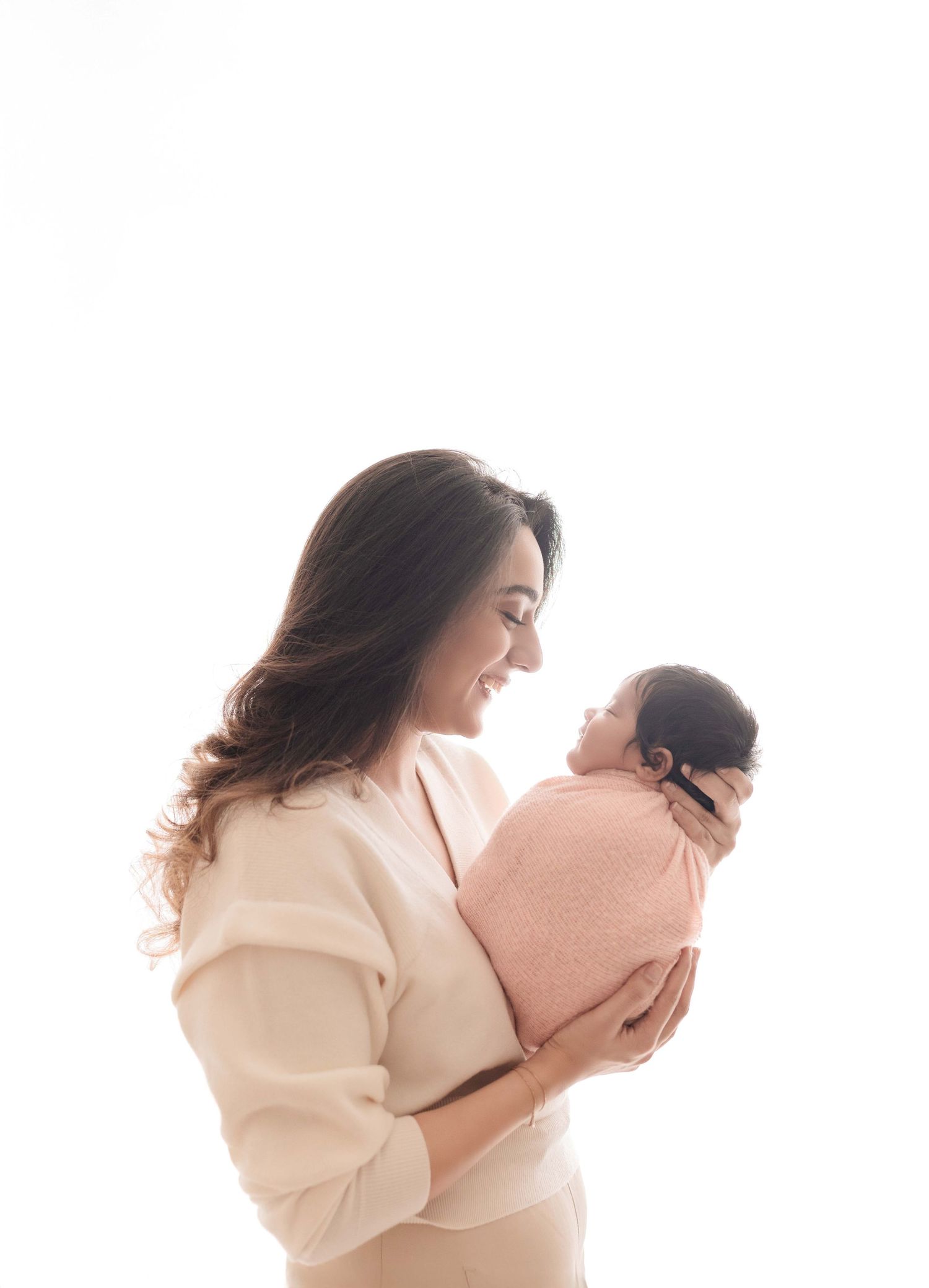 A smiling mother in a cream sweater tenderly holds her newborn baby wrapped in a pink blanket against a white background.