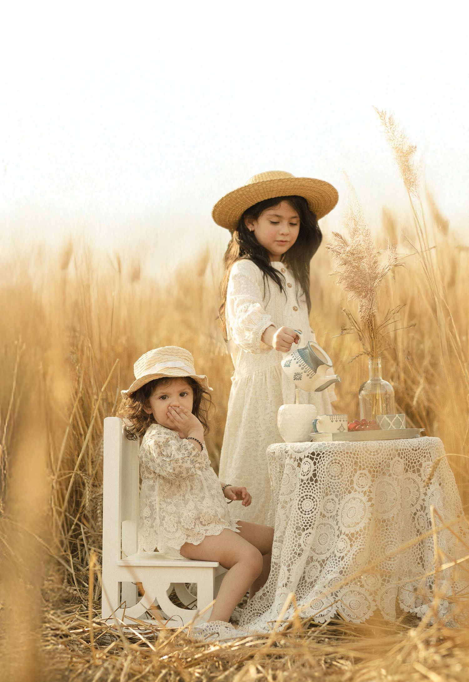 Two young girls in lace dresses and straw hats enjoy a tea party in a golden wheat field.