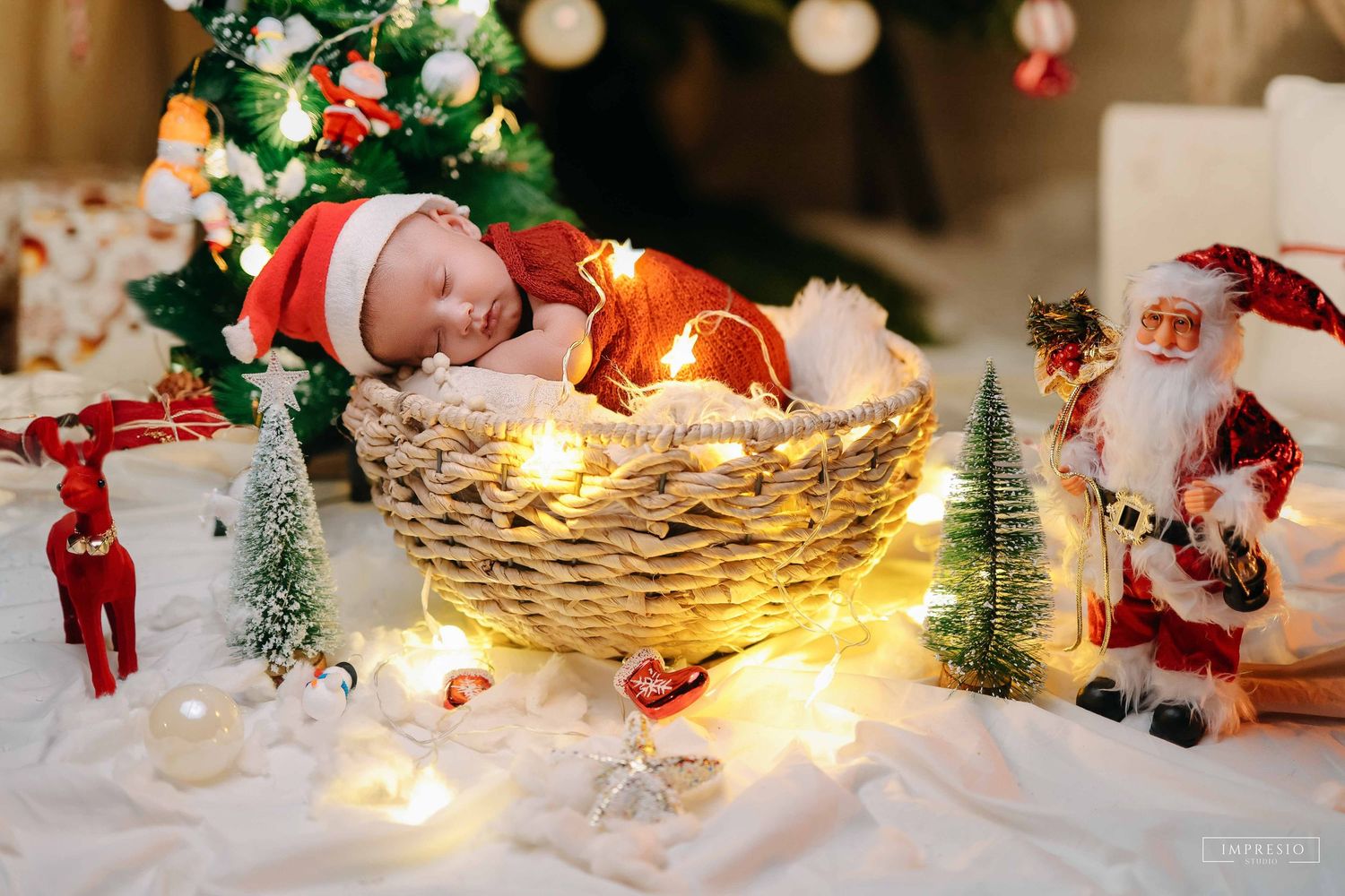 Newborn baby sleeping in a wicker basket surrounded by Christmas lights, Santa figurine, and festive holiday decorations.