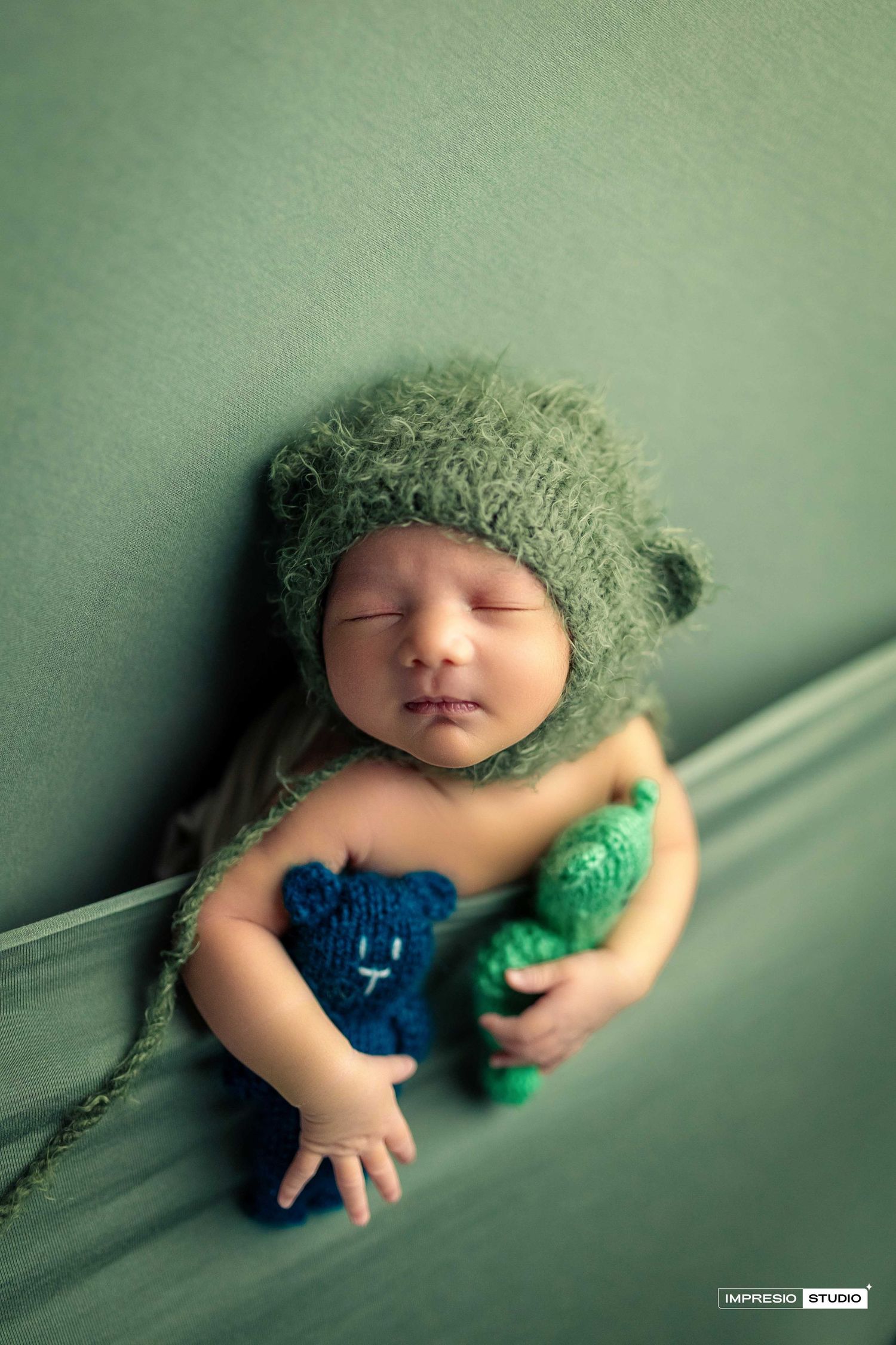 Sleeping newborn baby wearing a green knit bear hat, holding two small knitted teddy bears on a green backdrop.