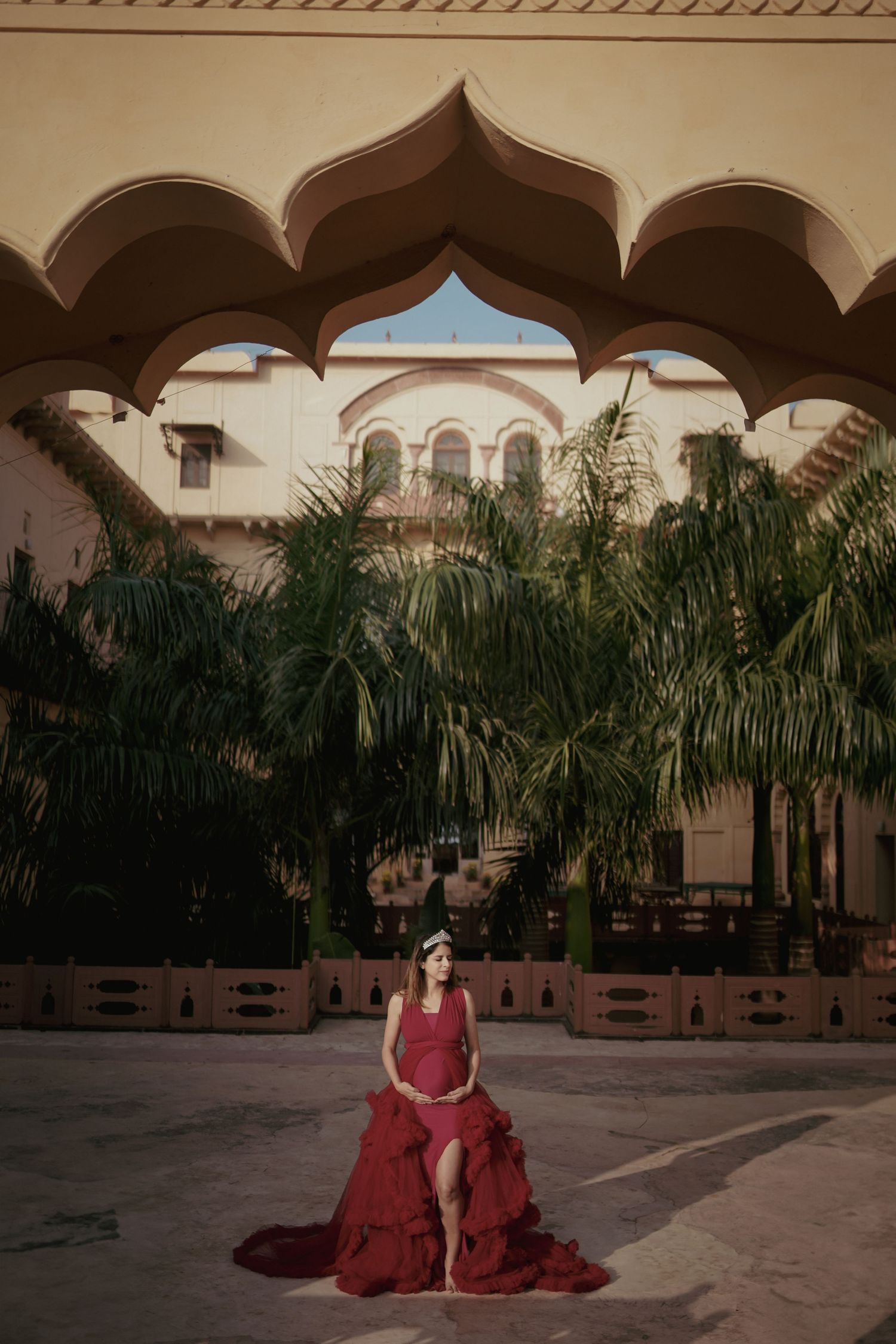Figure in a flowing burgundy dress poses elegantly in a Spanish colonial courtyard with palm trees for expecting mother photoshoot.
