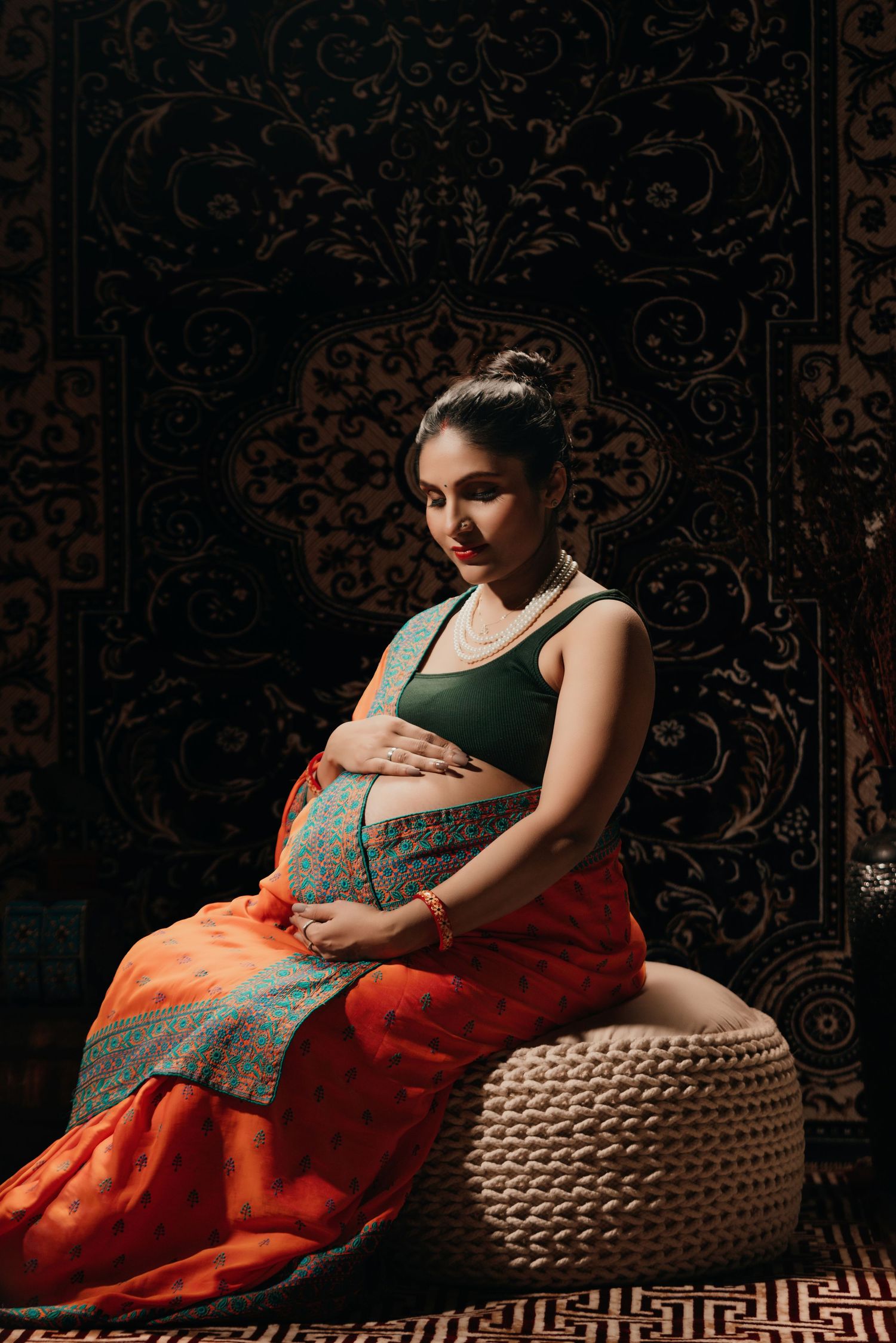 Artistic maternity photography with woman in orange and green attire reclining on woven ottoman with dark background.
