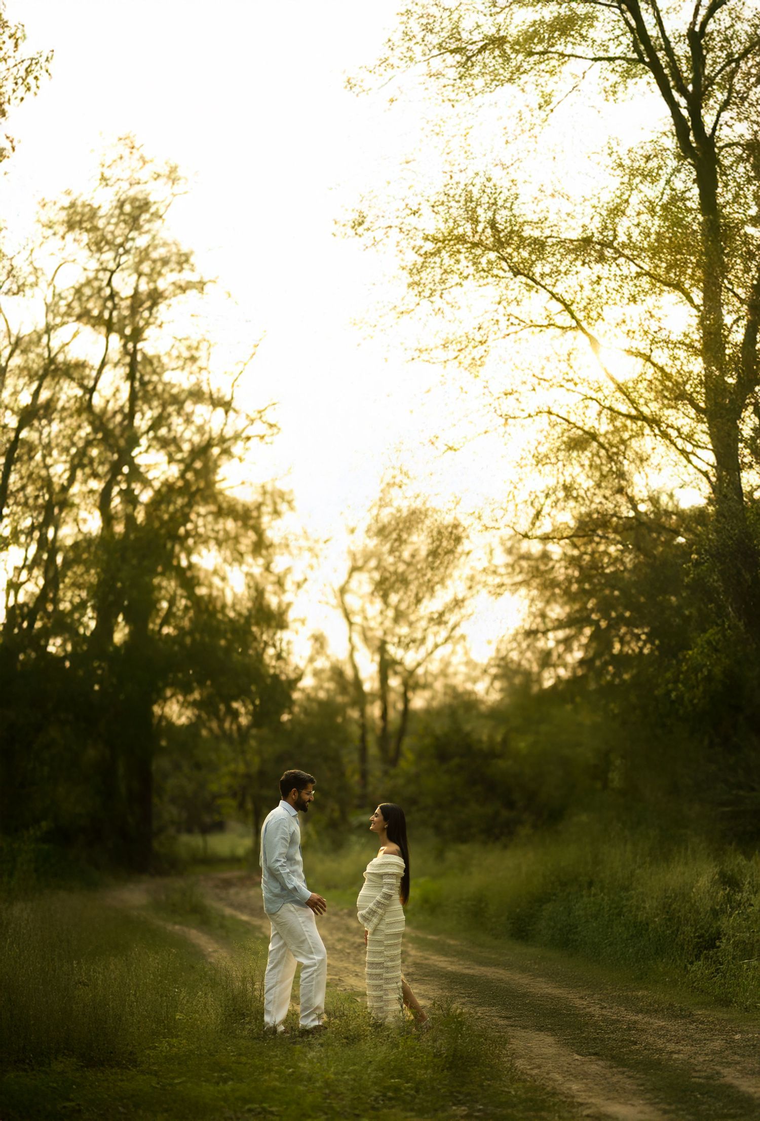 Two figures stand facing each other on a sunlit dirt path surrounded by trees at golden hour for outdoor maternity photography.