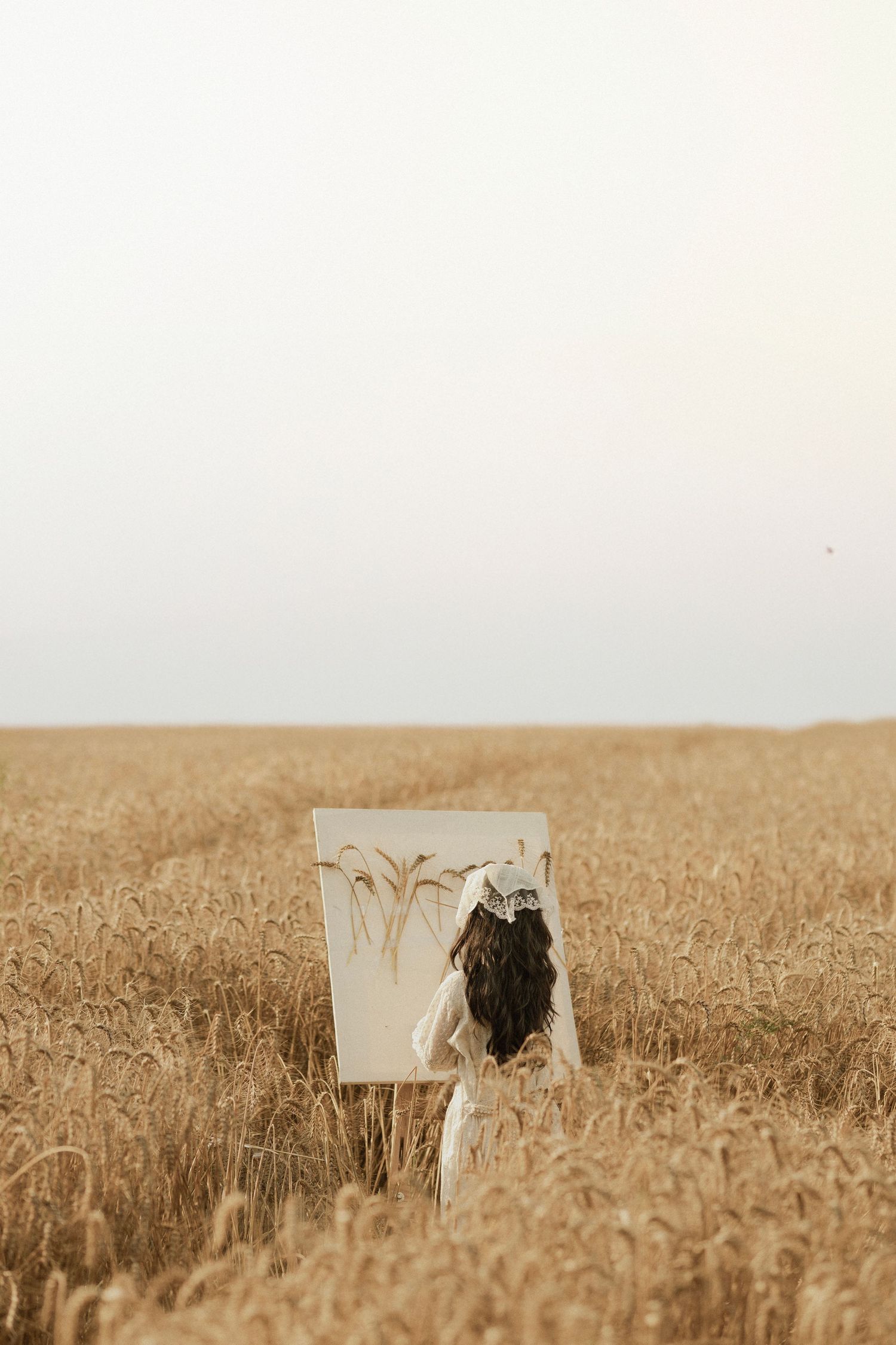 Woman in white dress painting on canvas in golden wheat field under overcast sky.