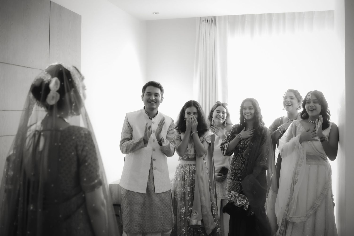 A black and white wedding photo showing a group of people reacting with joy and excitement in a bright interior space.