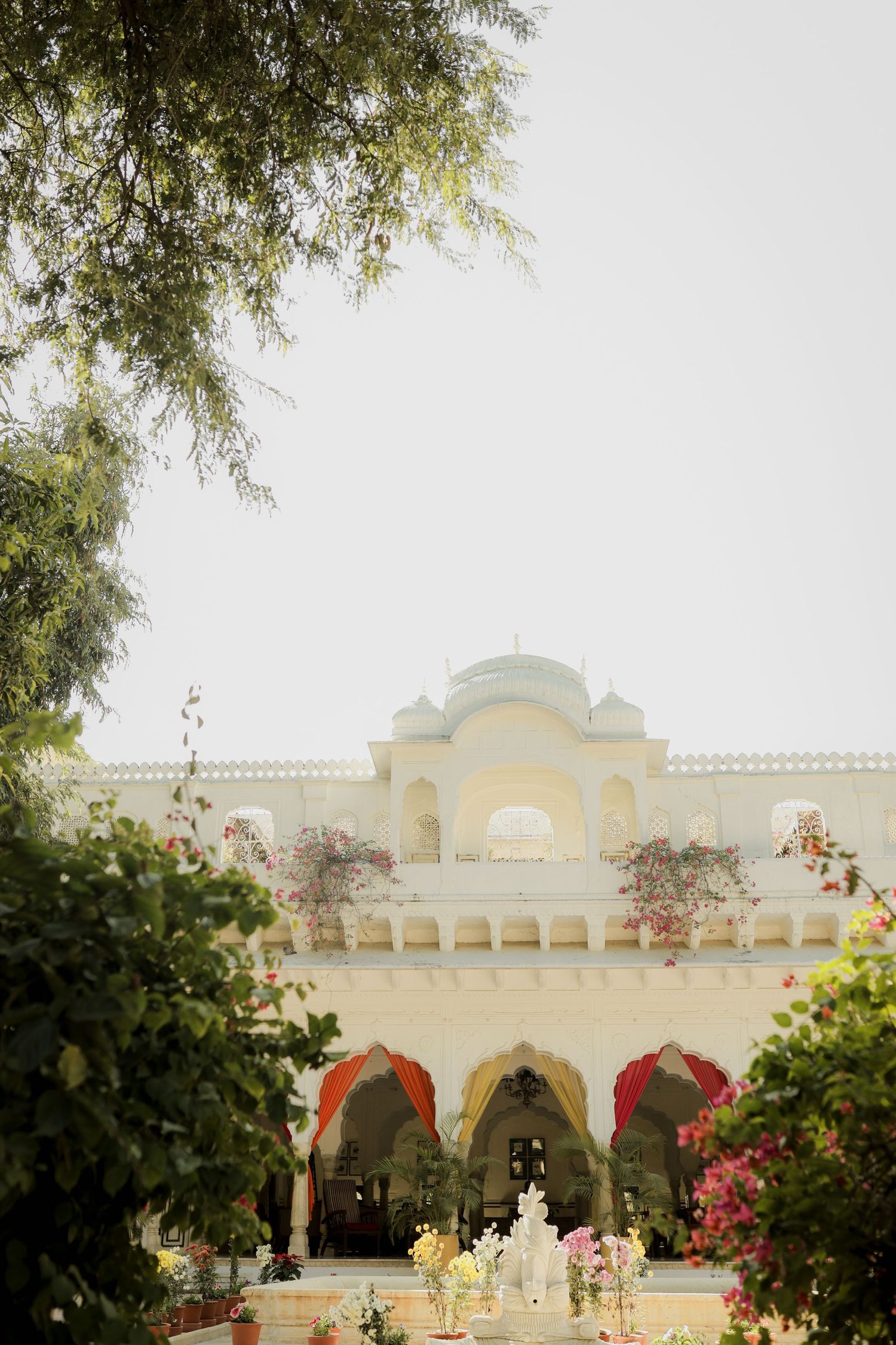 Luxurious white palace building with arched walkways and domes surrounded by lush gardens and flowering trees.