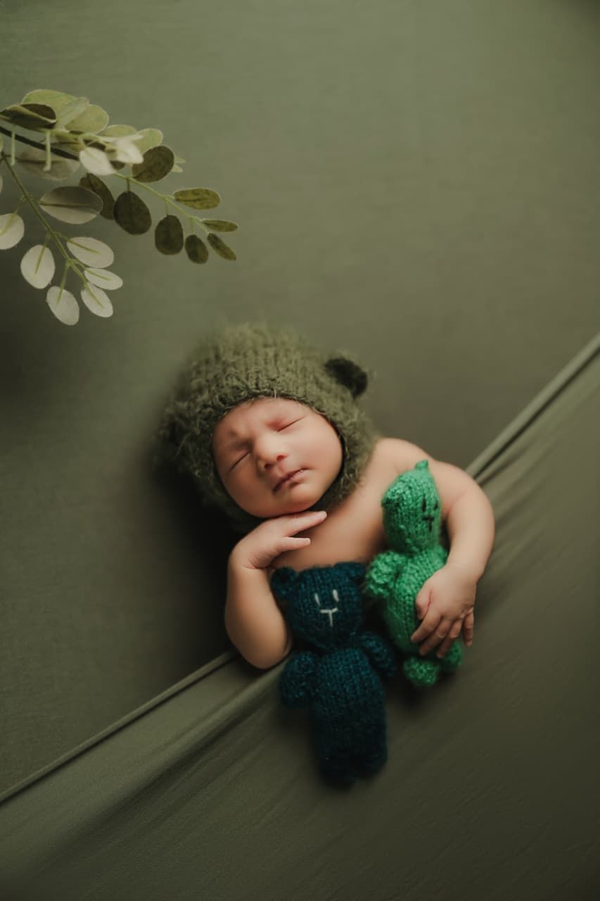 Sleeping newborn baby wearing a green knit bear hat, cuddling a teal crocheted bear toy on a green backdrop.