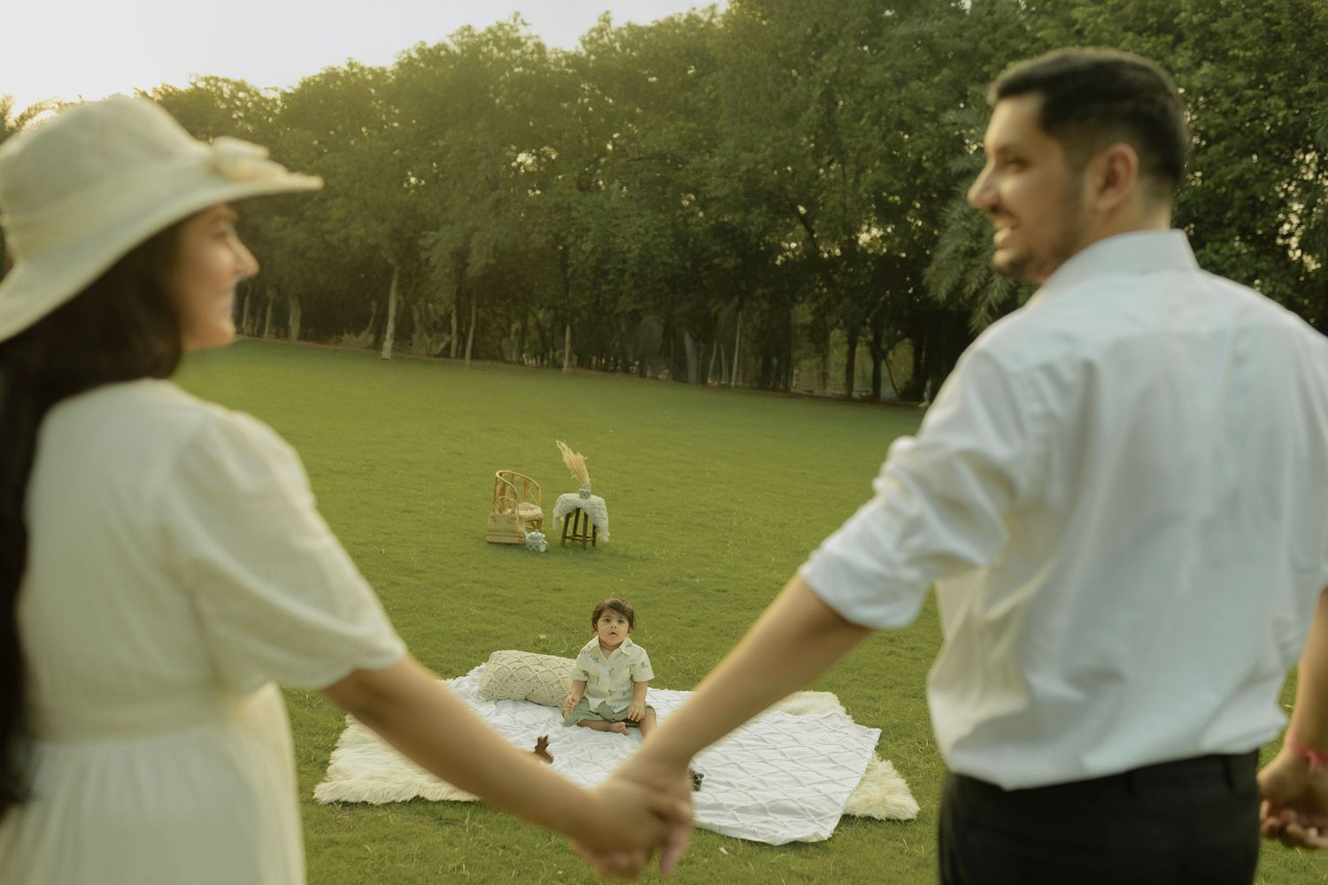 A couple holding hands at a romantic picnic setup on a green lawn with a sun-lit forest backdrop.