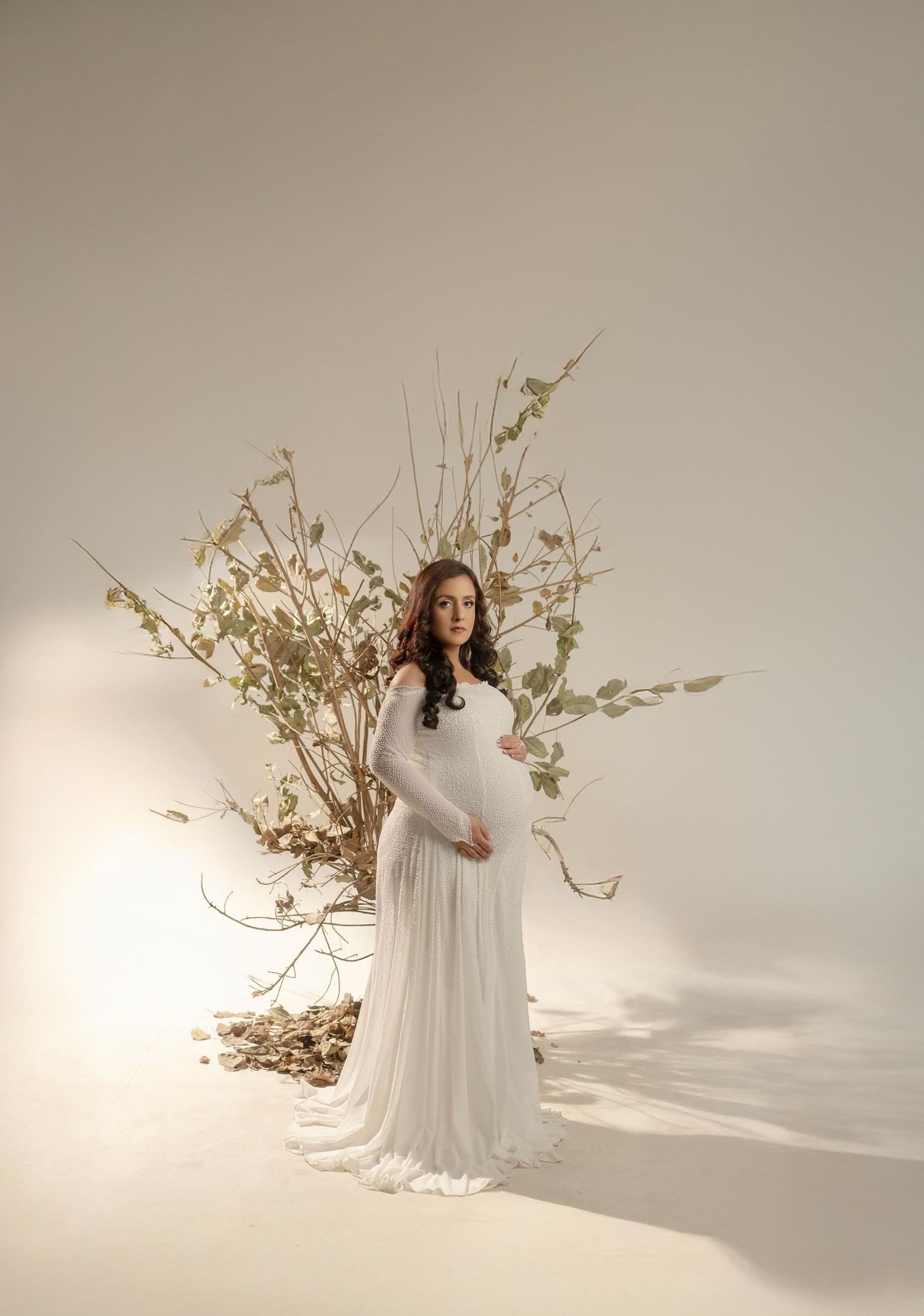 Graceful maternity session featuring white dress and natural branch decor in studio setting.