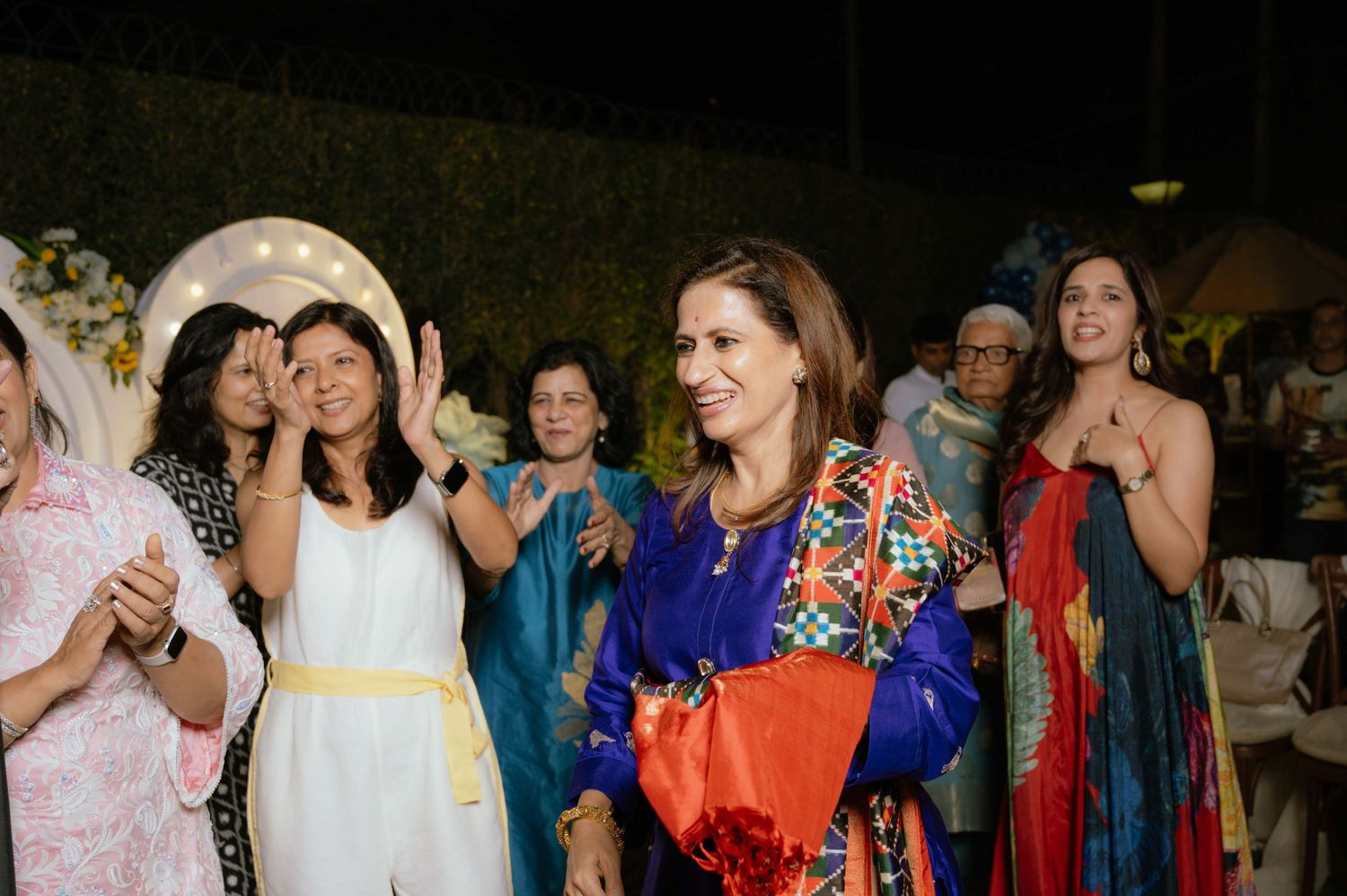 Women in colorful traditional attire celebrate at an outdoor evening party with floral decorations.