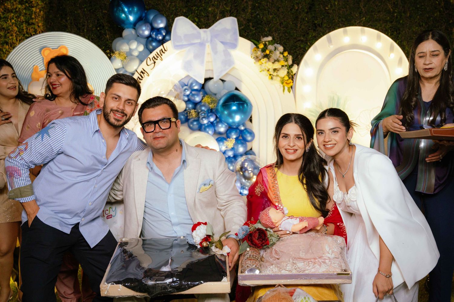 Group of five people smiling at a celebration party with blue balloon decorations and a cake.