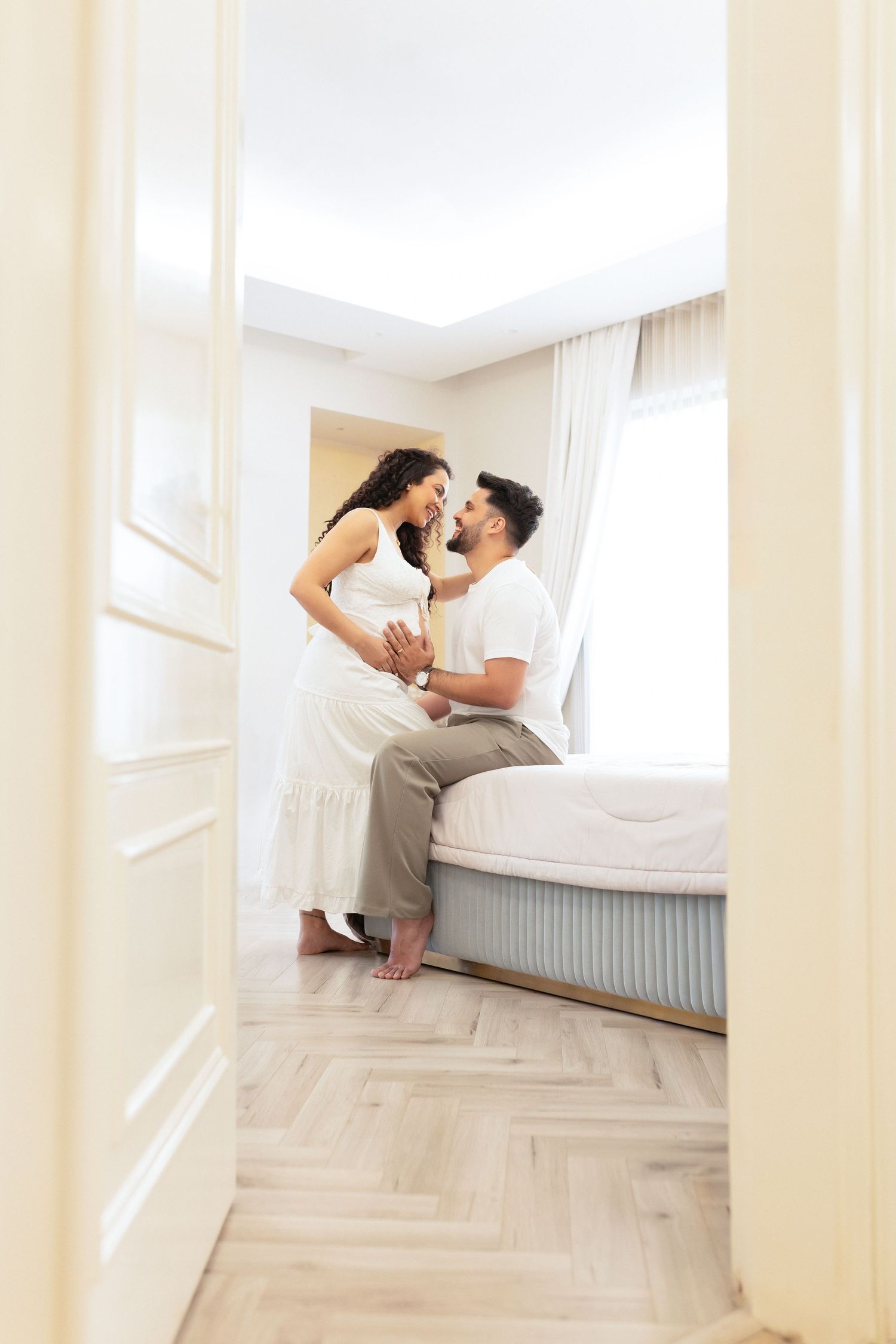 A couple playfully jumps on a mattress in a bright white room with hardwood floors.