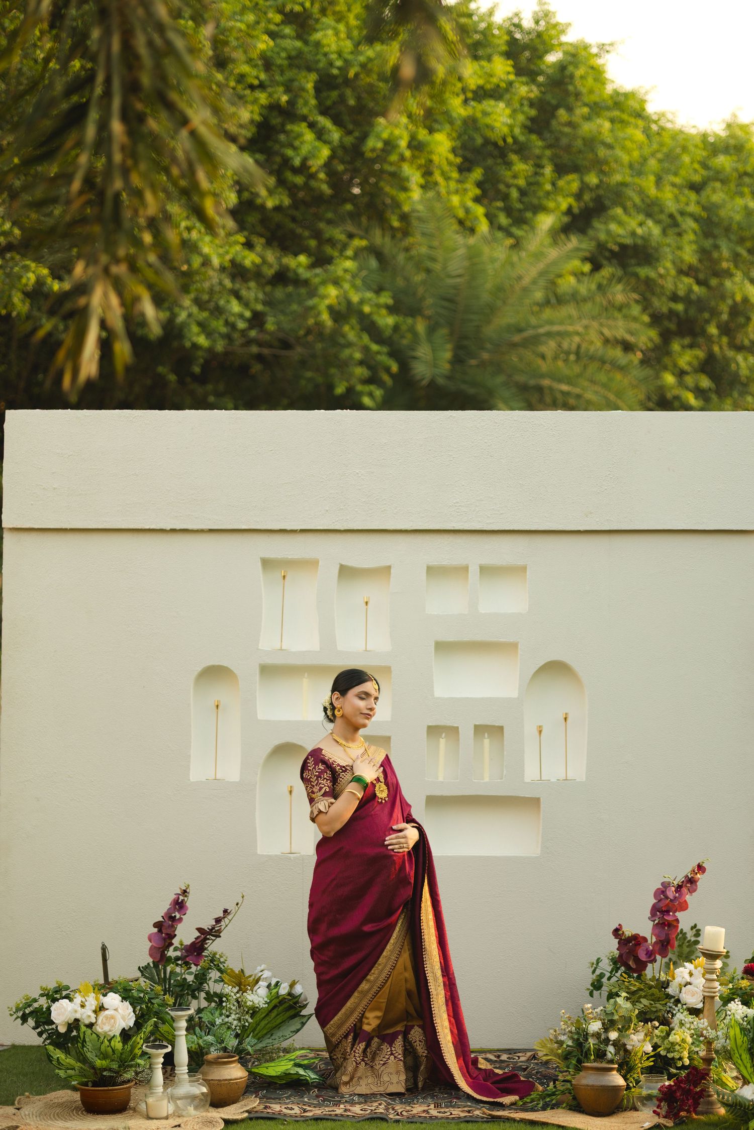 A person in a burgundy and gold sari poses elegantly against a white wall surrounded by potted plants and flowers.