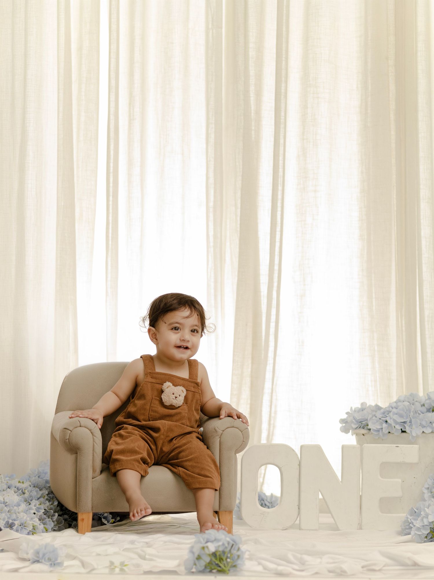 Smiling baby in brown overalls sits on a mini chair beside ONE letters and blue flowers for a first birthday photo.