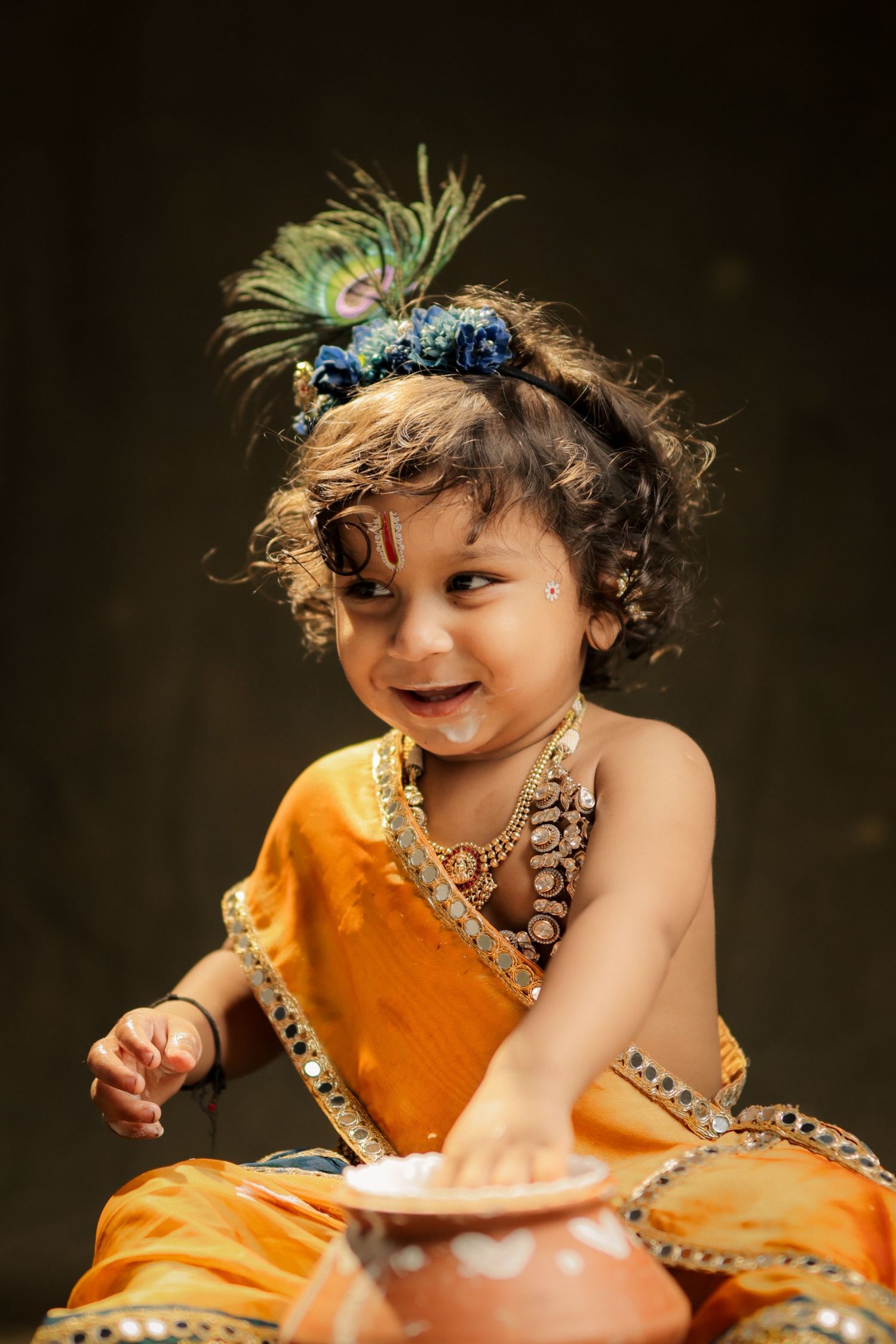 Joyful toddler dressed as Lord Krishna in orange attire with peacock feather crown, smiling with white cream on face.