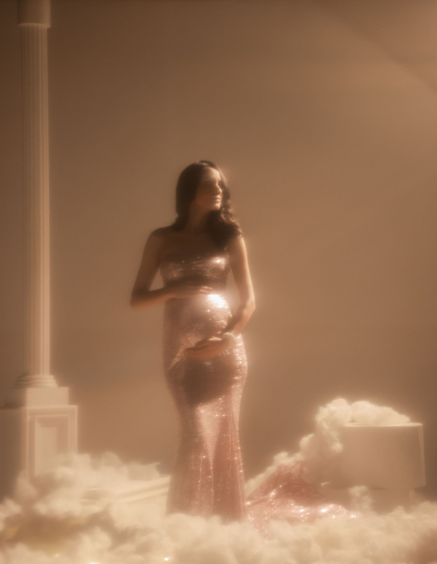 Pregnant woman in a sequined rose gold gown cradling her belly, surrounded by soft clouds and warm golden light.