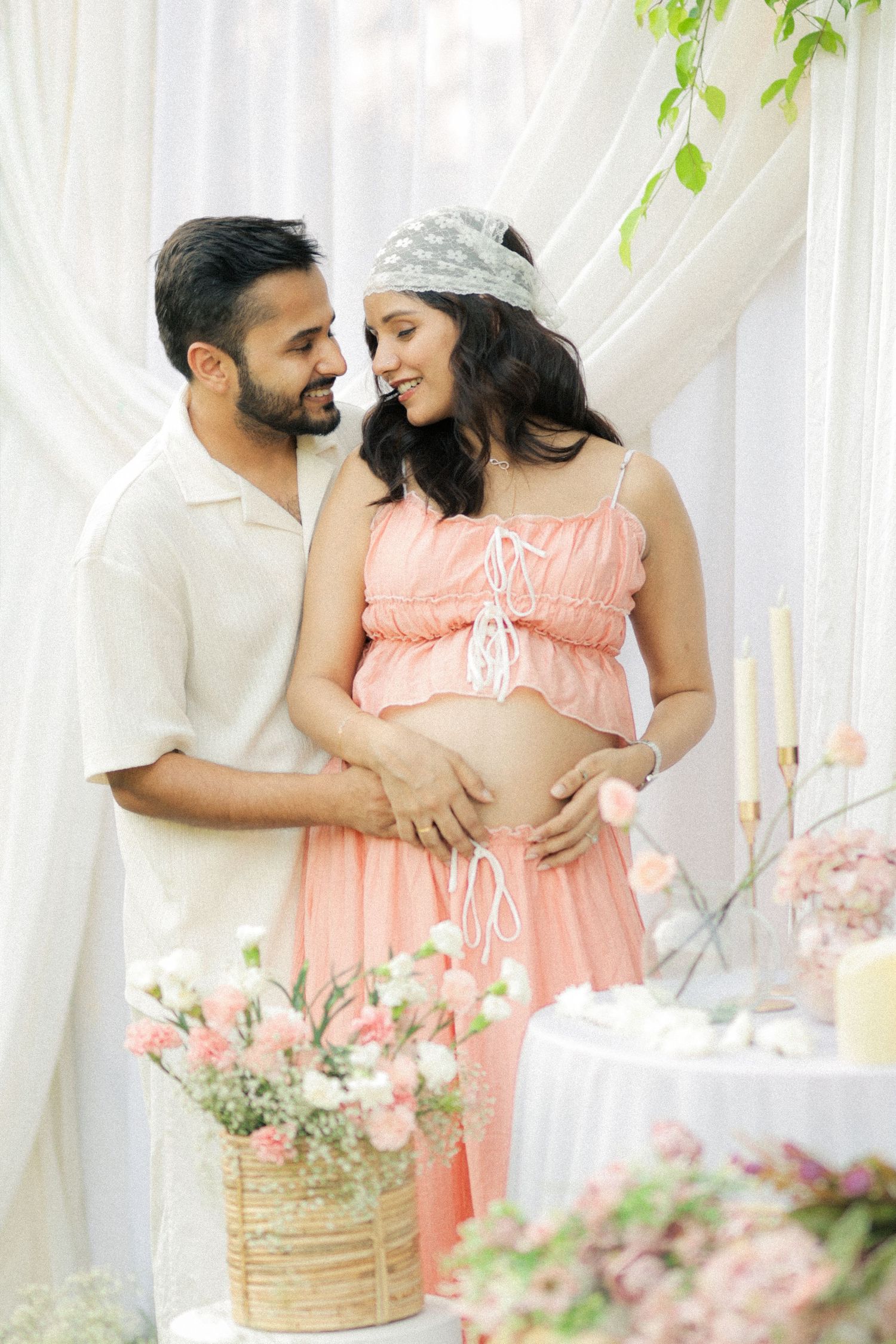 Happy couple at baby shower, pregnant woman in pink crop top and skirt, man in white shirt, floral decorations.